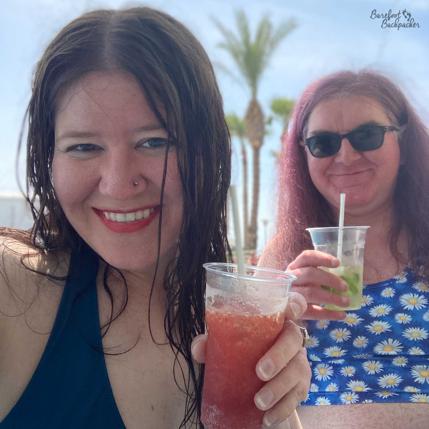Two people take up most of the shot. They are both holding cocktails. The person on the left is a woman with long straight damp hair and a nose-ring. The person on the right (an enby) is wearing sunglasses, a daisy-motif crop-top, and long purple-ish hair.