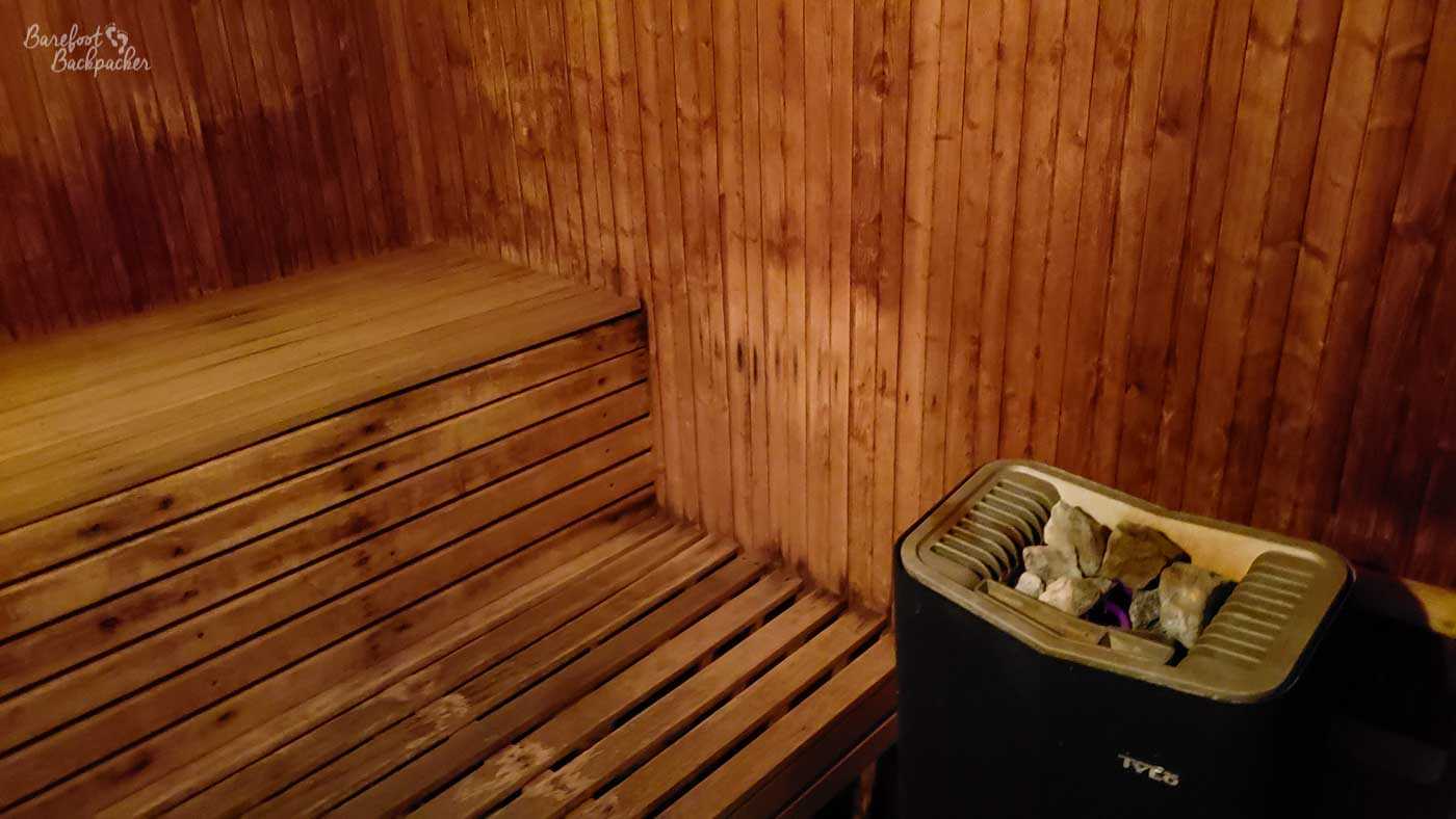 Image of the inside of the sauna. It's a vertical slatted wooden wall, with a horizontal slatted wooden bench fixed to the right side. In front of the bench, on the wall, is a heater with coal at the top.