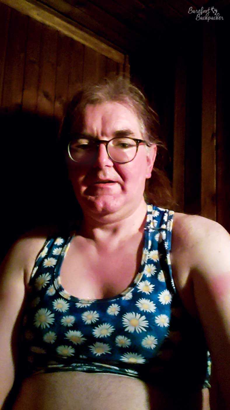 Someone is sat in a dark room wearing a daisy-motif crop-top, and glasses. They look quite sweaty/hot.