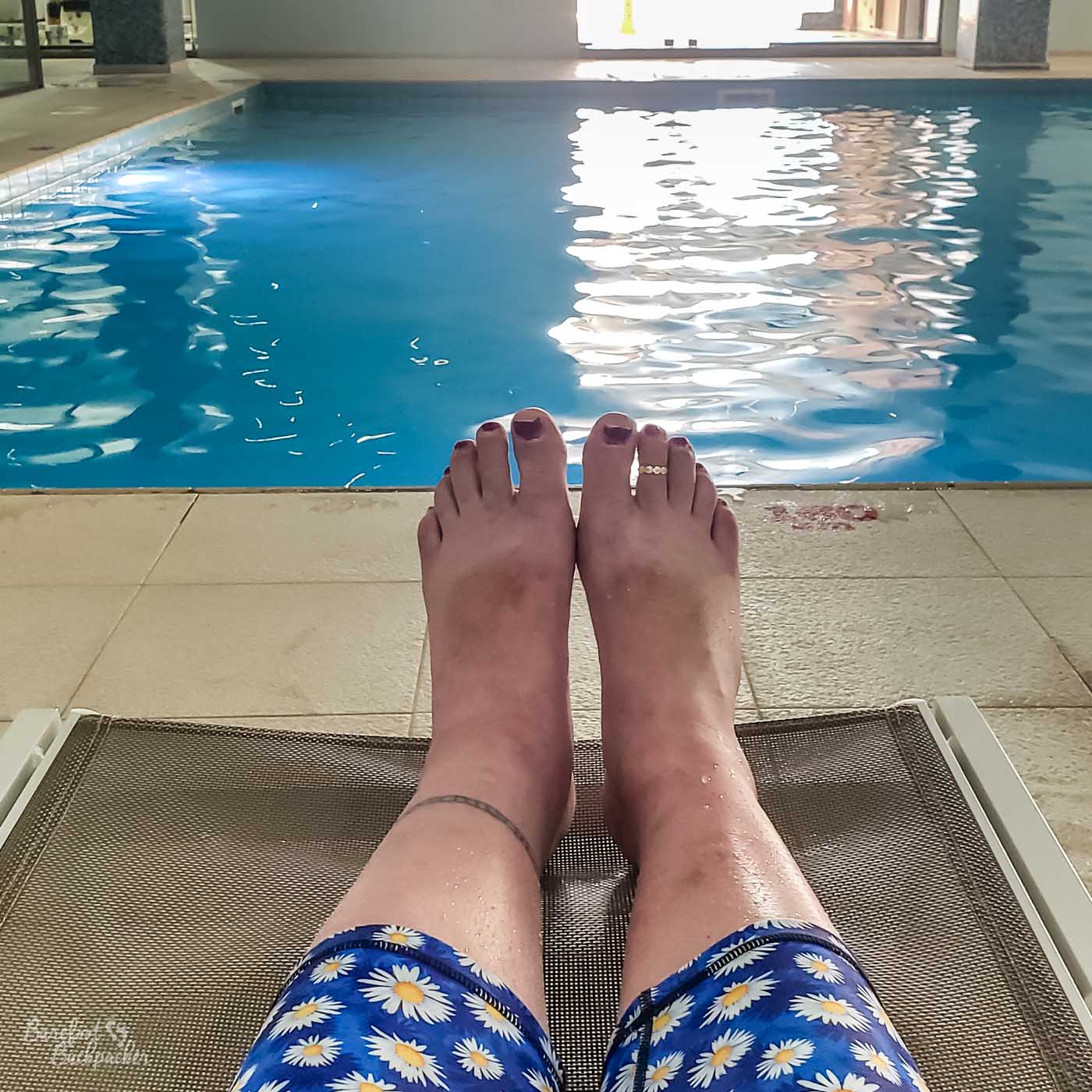 Lower legs (partly covered in daisy print leggings) and feet (bare aside from purple painted toenails) are reclining on a mesh seat; there is an indoor pool in the background.