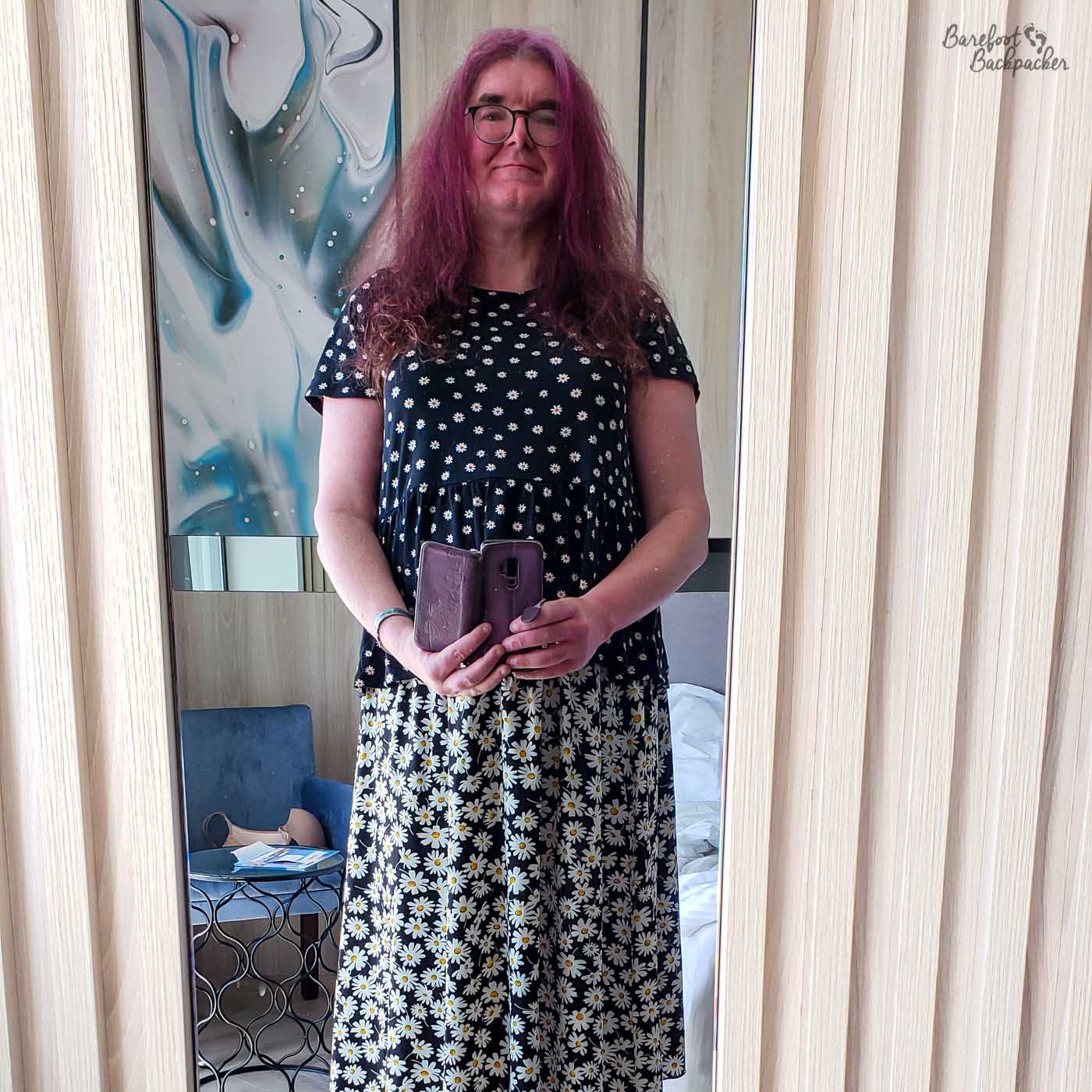 Person is taking a selfie in front of a mirror. They are wearing a daisy-print blouse and a daisy-print skirt, which don't match patterns. They have long straight hair and are holding their phone down and out in front of them. The picture is taken in a hotel room.