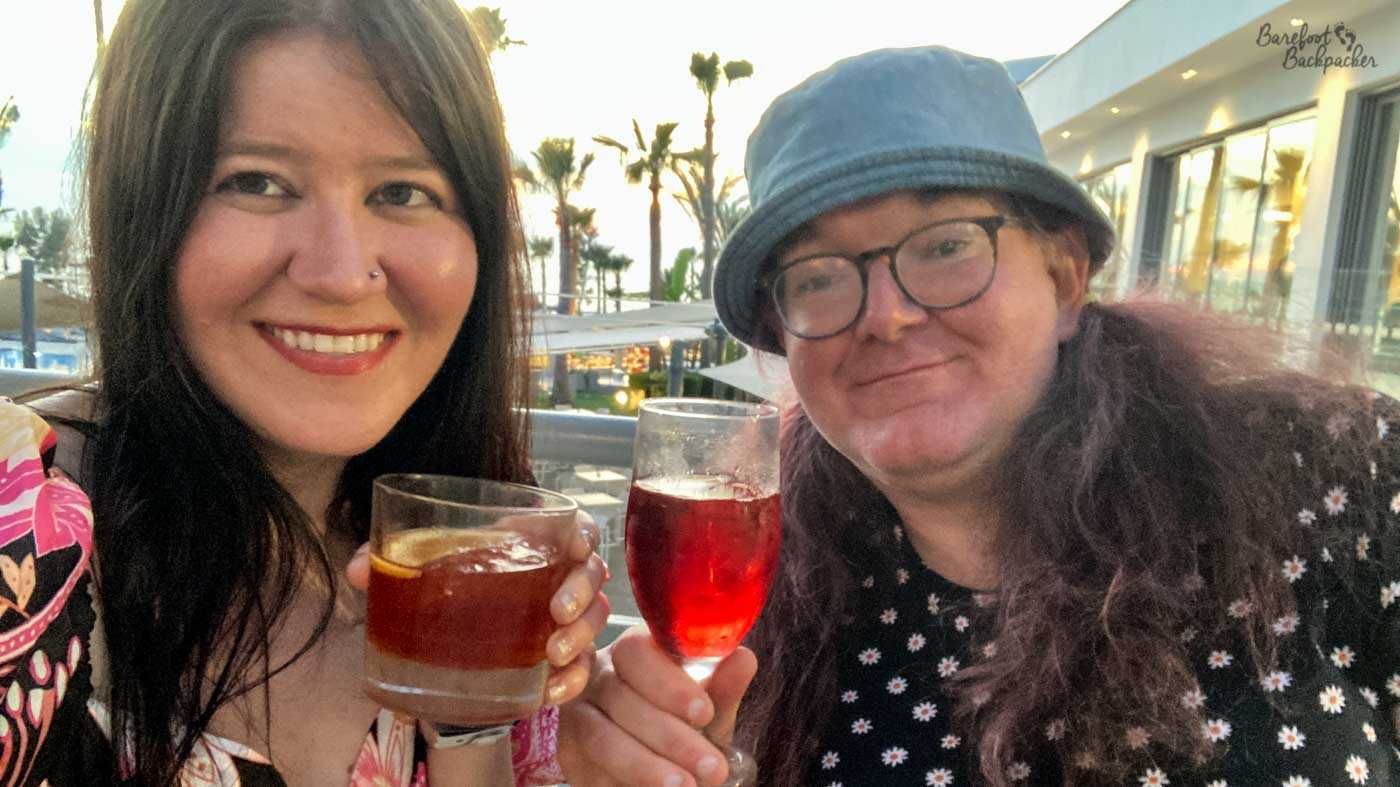 Two people smiling awkwardly at the camera, sat down in front of a glass panel fence through which down below there's a patio area. The woman on the left is holding a whisky-type glass. The enby on the right is holding a wine glass.
