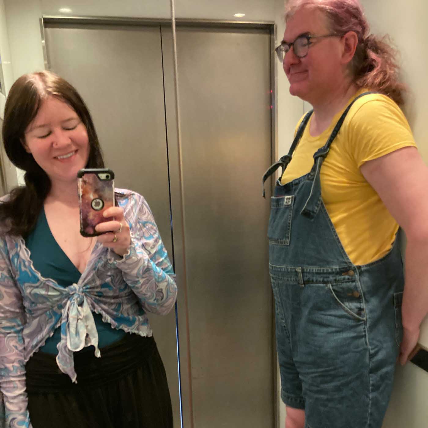 Two people are in a lift (elevator). The woman on the left is taking the picture with her phone, using the mirrored walls of the lift as a reflective screen. She has long straight hair, a fabric tie-top over a t-shirty type thing, and a skirt. She is smiling. The enby on the right is standing against the wall of the lift, taller than the woman so their gaze is over her head. They are wearing a yellow t-shirt and blue dungaree shorts, making them look a bit like a Minion.