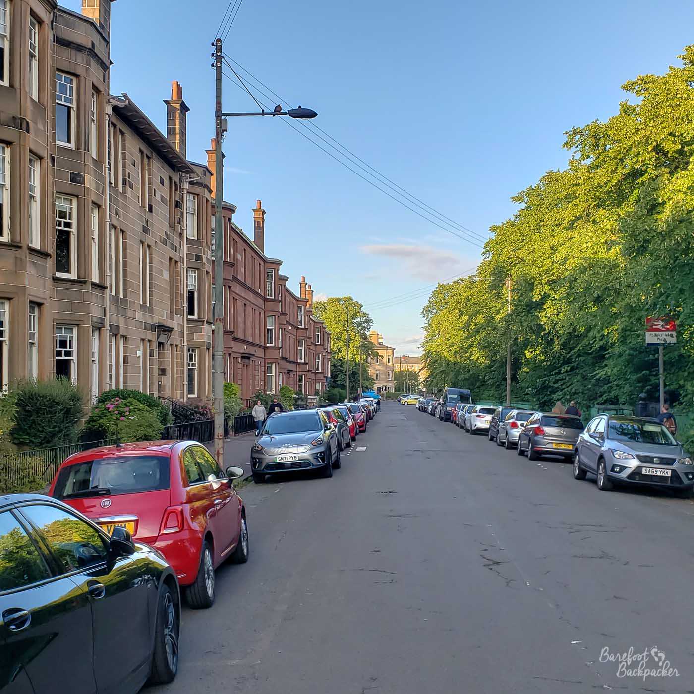 View of a street. On the left of shot is a row of tenement block. On the right are bushes and trees. In the distance is a railway station sign - Pollokshields West.