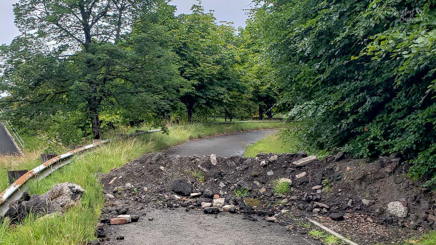 A narrow road bends to the right, with crash barriers on the left and trees on the right. In the middle of the road is a defined pile of rubble, small enough to easily climb over but too big to drive over.
