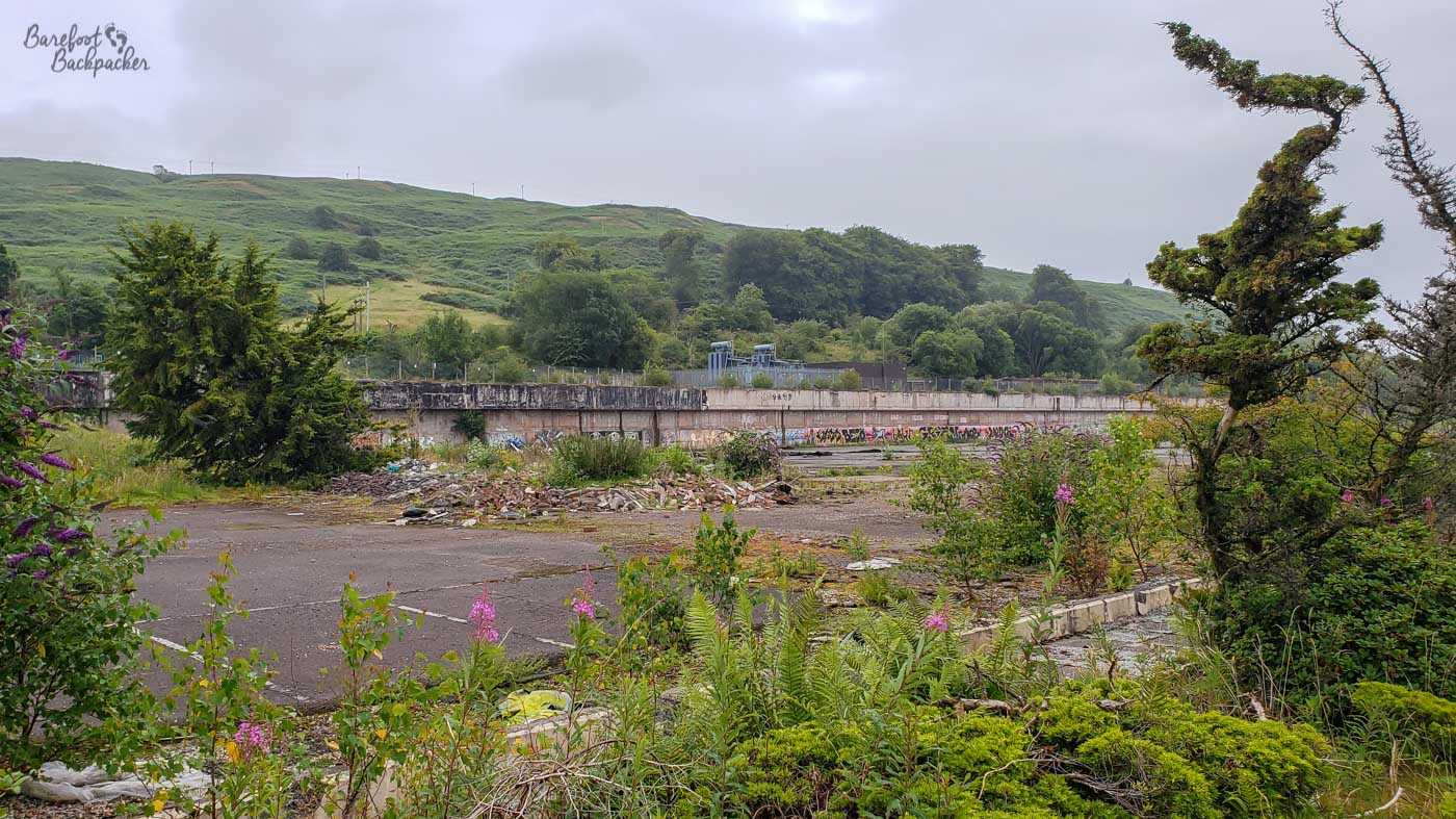 A car park covered with weeds and flowering heather. Rubble hides some of the demarcation lines. There's a long concrete wall on the far side of the tarmac, behind which are steep hills.