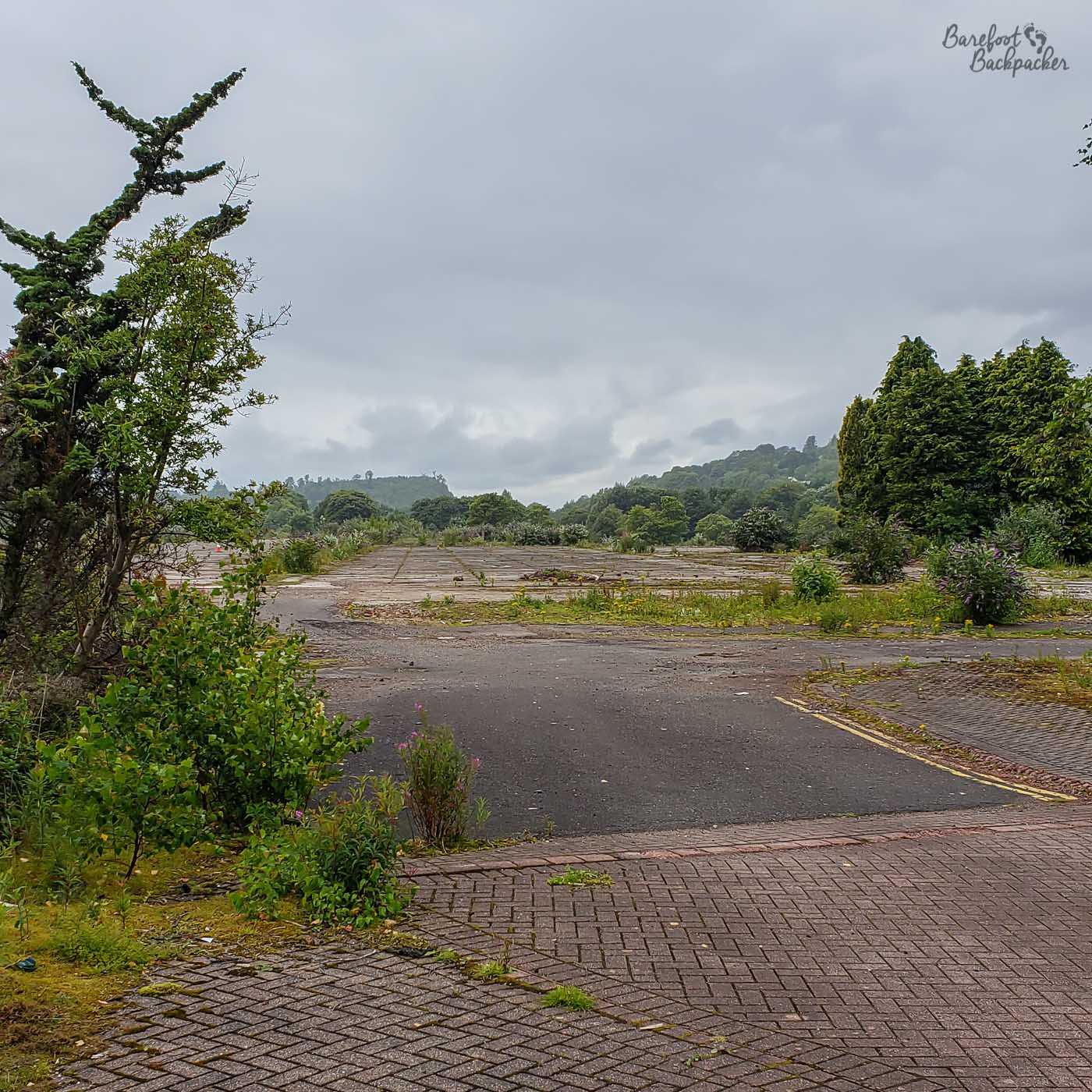 Another wide open space lined with concrete that used to be a building. The road leading to it can clearly be seen, and the site itself has shrubs around the edge marking where the walls used to be.
