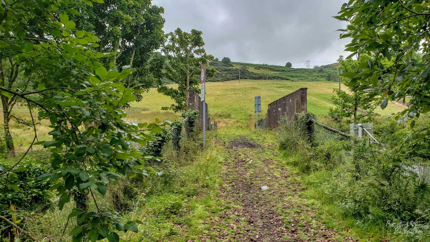 A small roadway surfaced with stone and gravel narrows as it goes over a small brick bridge. There's a signpost pointing in the wrong direction that warns it's a weak bridge. To the right, front of shot, are fences that mark where access to the platform used to be. Beyond the bridge is hill, grass, and farmland.