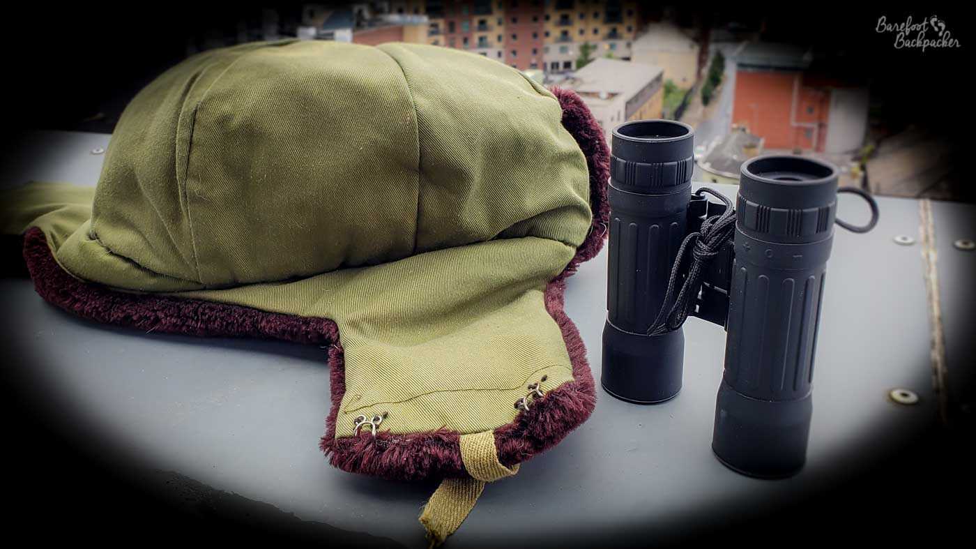 Image of a furry hat with ear-coverings and a pair of binoculars lying on an apartment's balcony wall. The sides of the image are darkened giving a vignette effect.