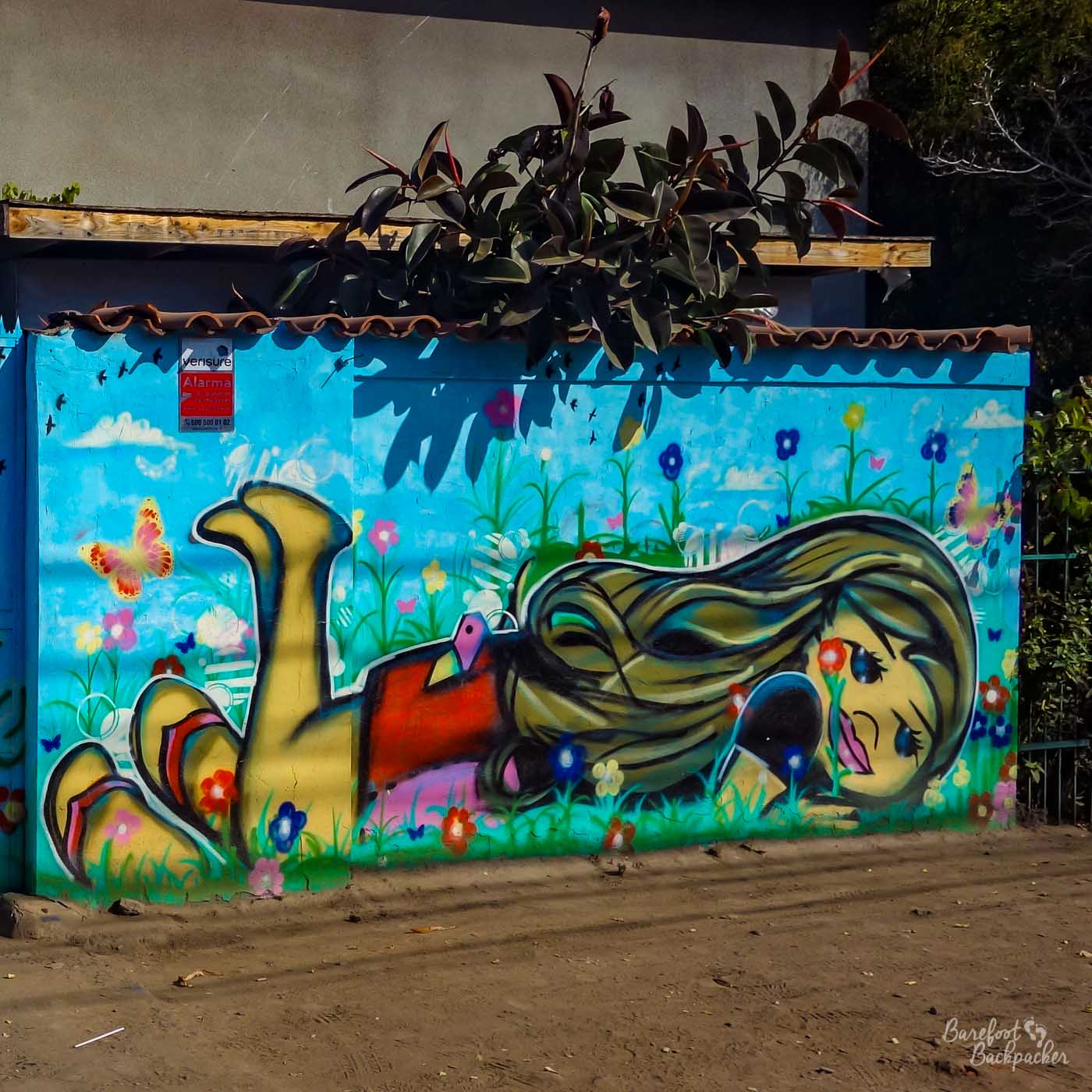 A piece of colourful street art on a wall. It's of a girl with long hair, lying in the flowers on her stomach, with her legs bent up in the air. Her sandals are on the grass behind her.