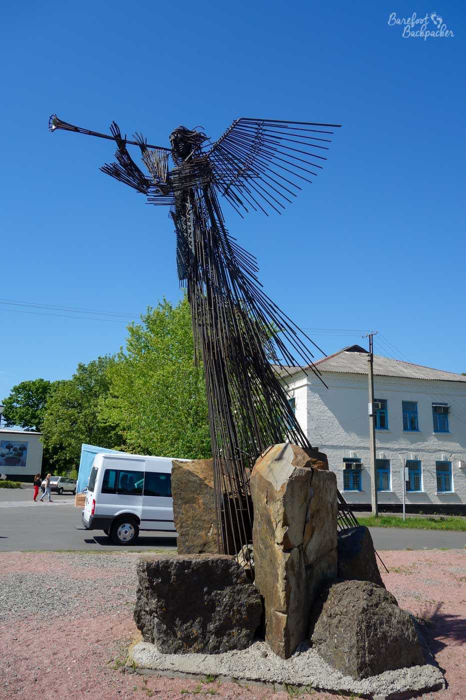 Standing in a pile of boulders is a statue made up of little pipes of wire. It's a tall statue – several metres in height, and it's of a figure with a long cape and wings, blowing a long straight horn-type instrument. It is dark against the bright blue sky and green trees behind it.