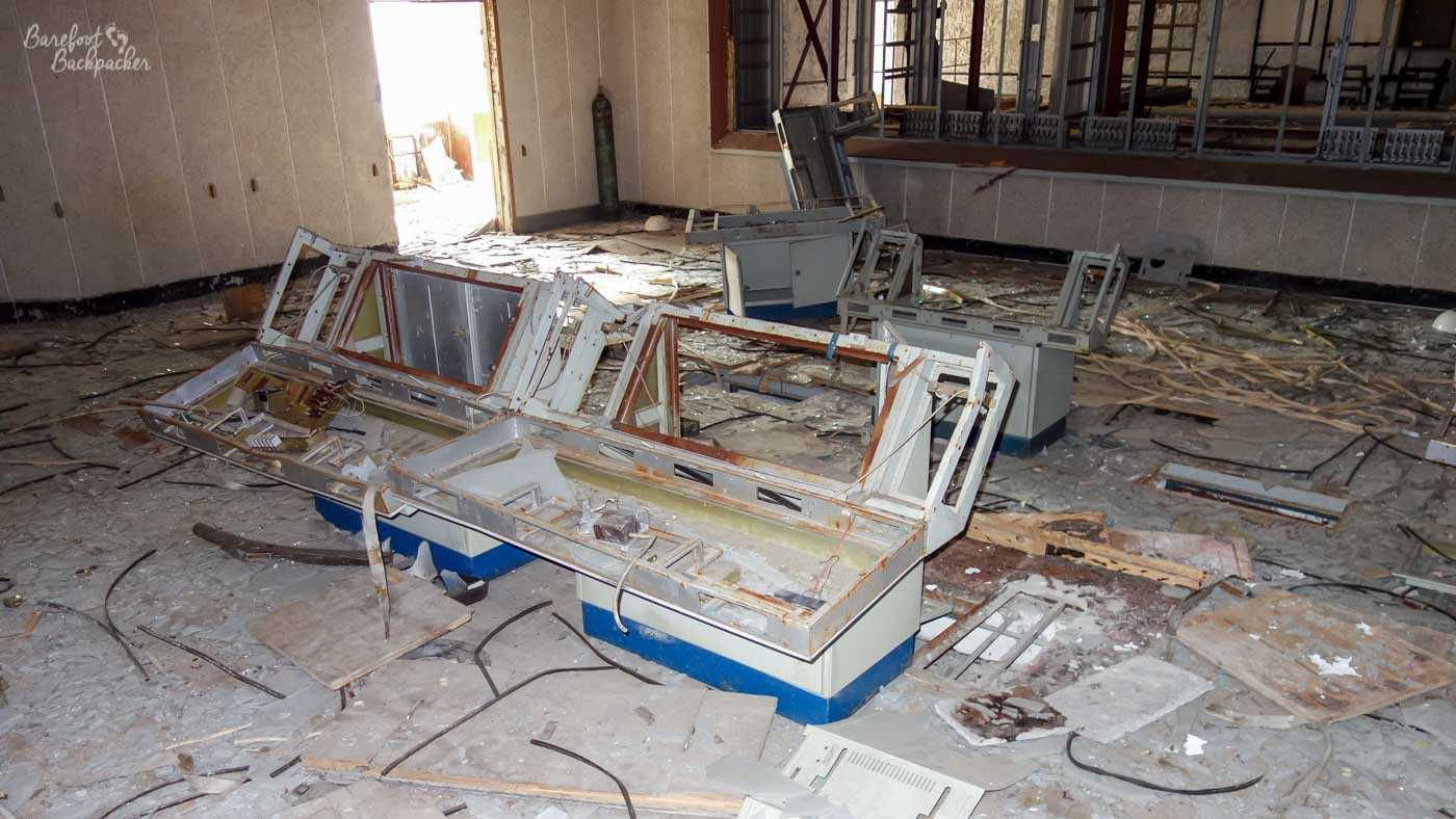 A large open-plan room with a broken desk console of some kind of the centre. Glass, wood, wires, and paperwork are scattered on the floor nearby.