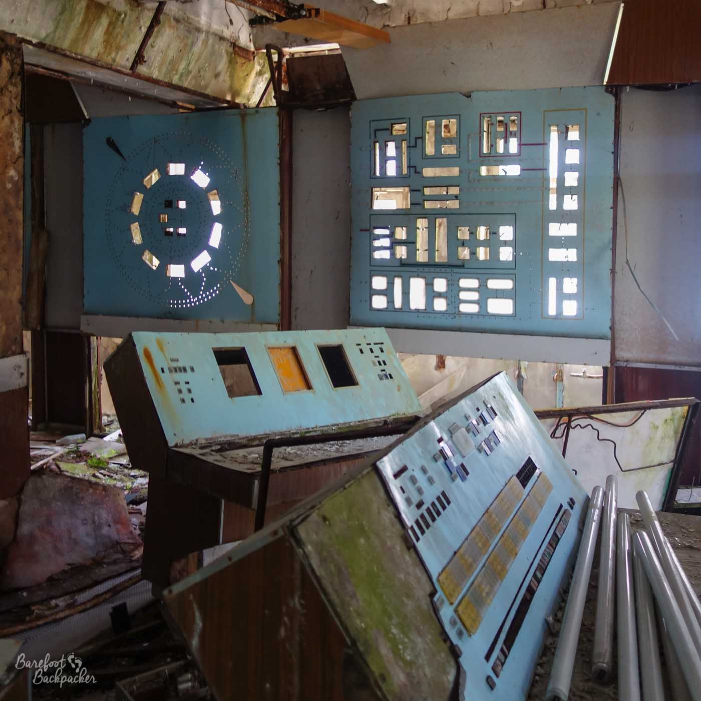 Picture of a broken and derelict control room at the Duga Array near Chernobyl. In the foreground are two control desks, with holes where buttons used to be. On the wall are two broken panels where display lights would have been.