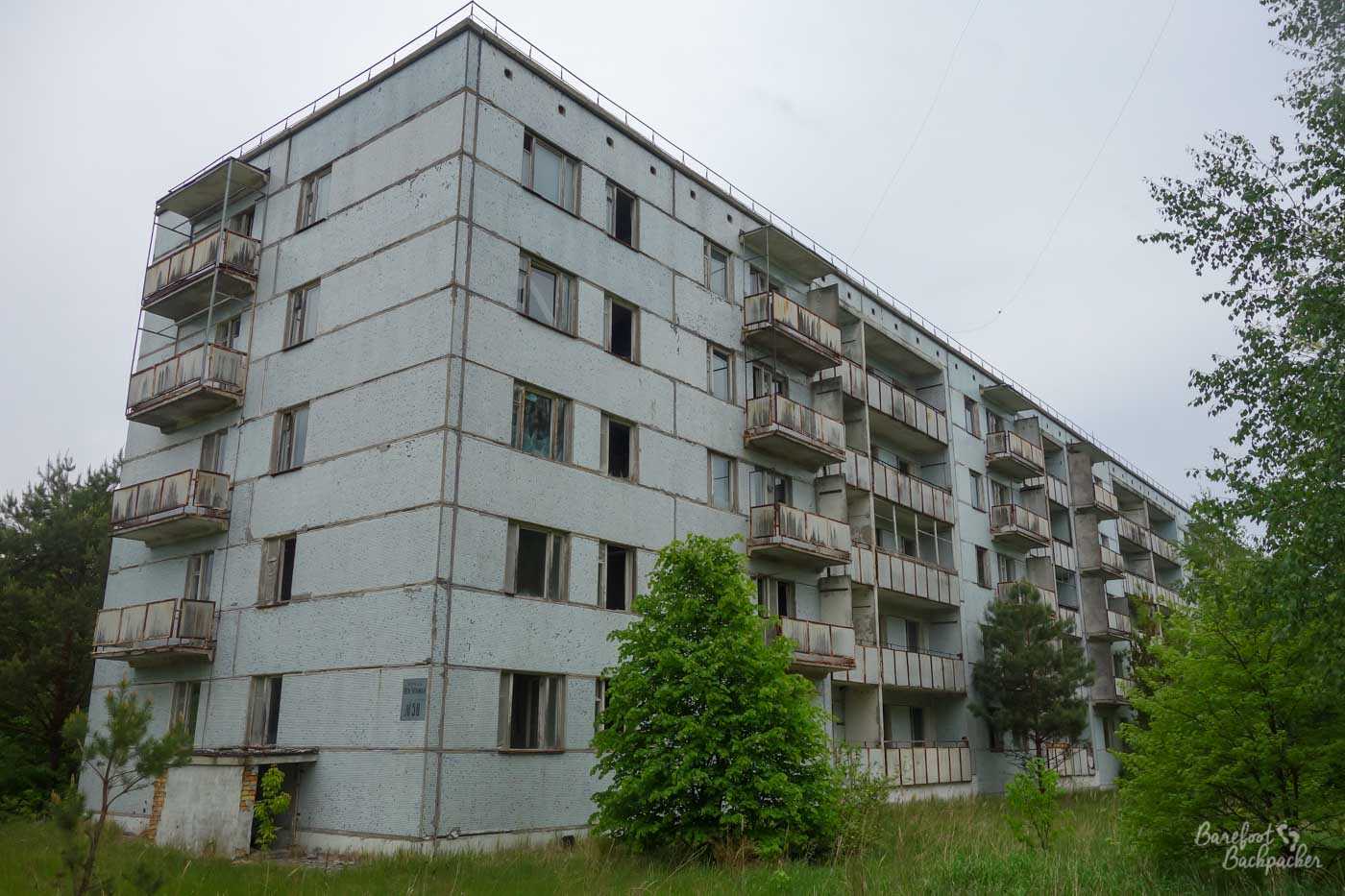 One of the many apartment blocks in Pripyat. This one is five storeys high, quite long, and with a very plain white exterior. Each flat has a small balcony. There are trees growing very close and slightly up it, but not through the walls, yet.