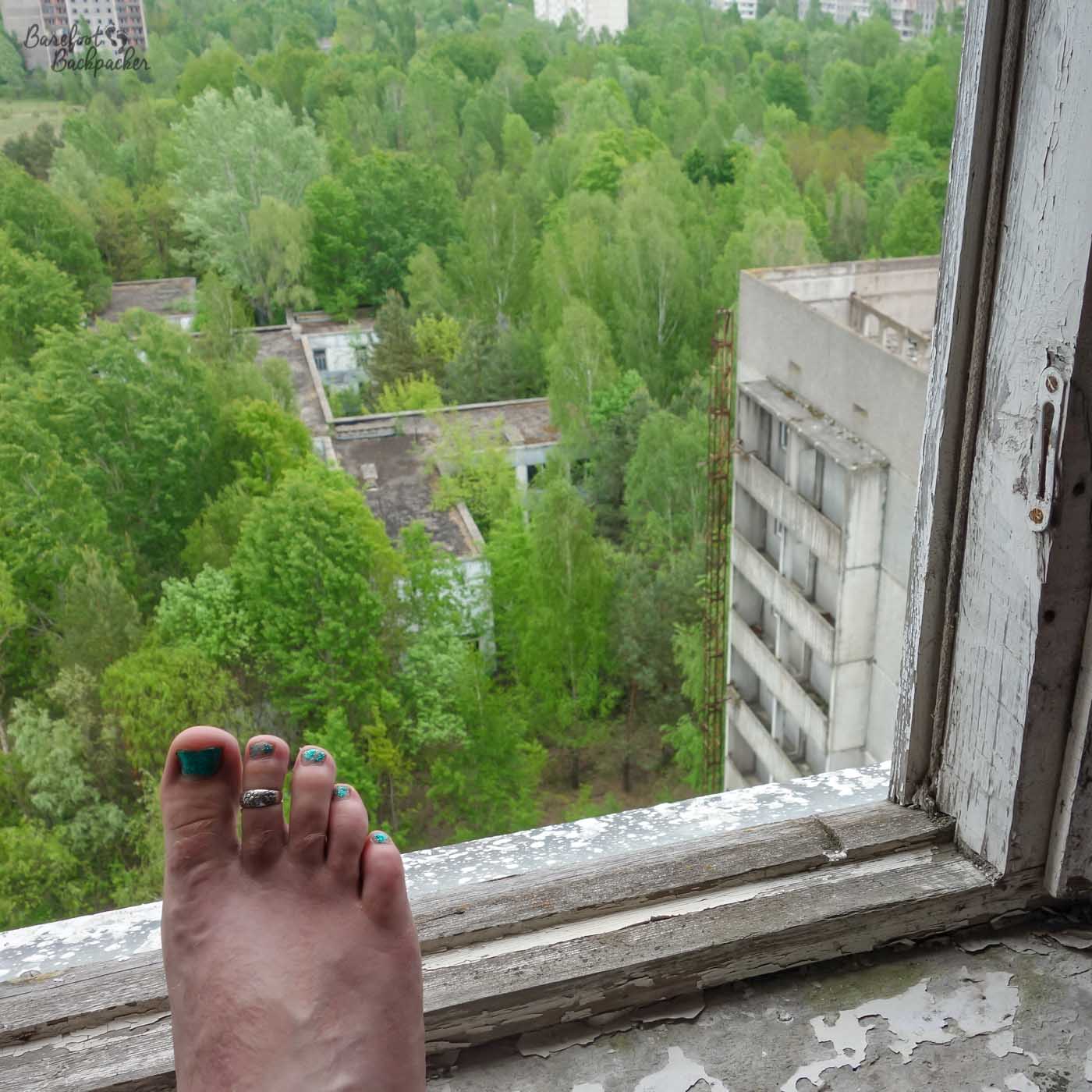 Picture taken from inside one of the apartments in Pripyat, looking out through a window. Outside are trees. In front of the window is one bare foot.