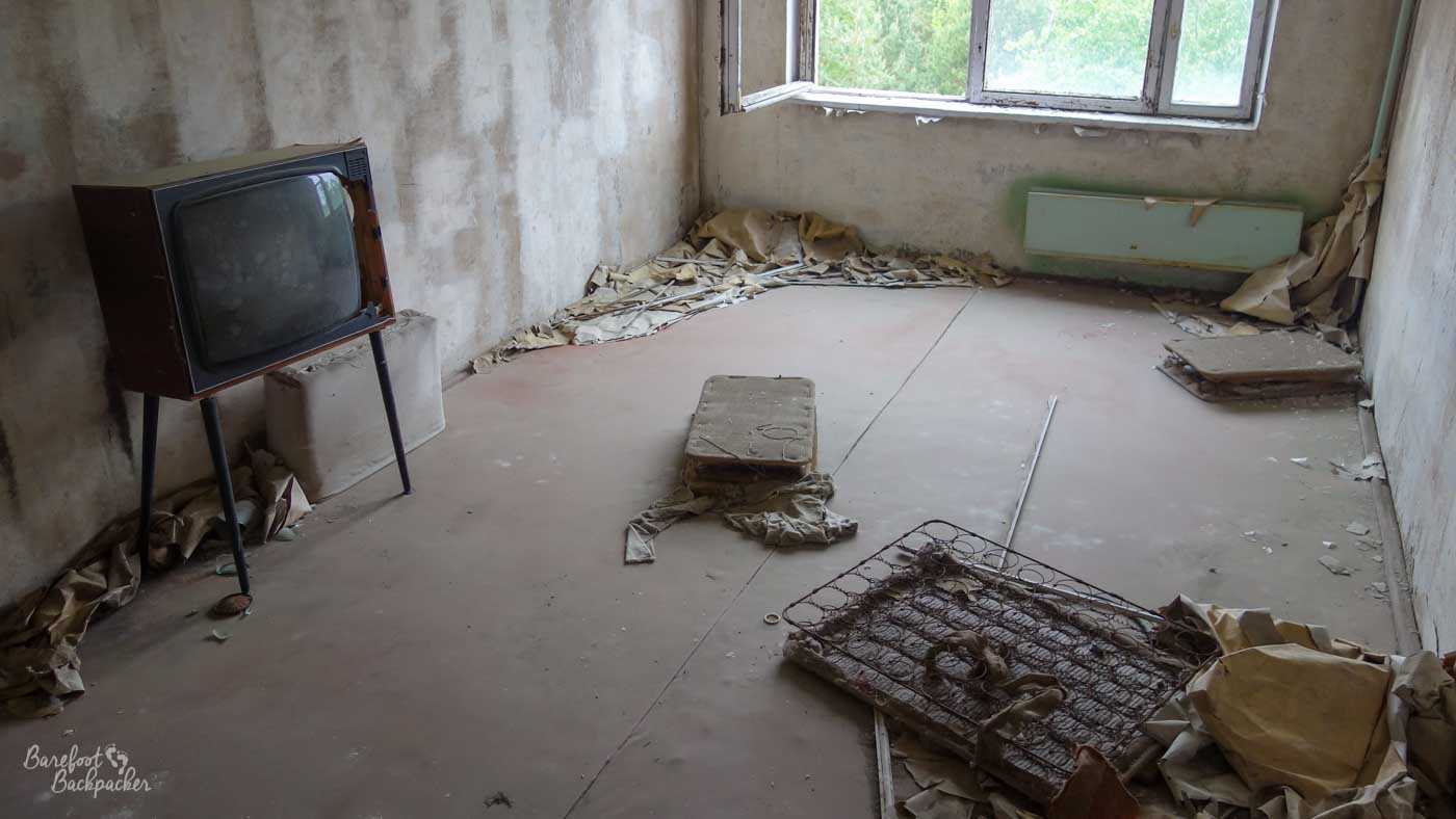 Inside one of the apartments. It's a rectangular room, plain walls, large window on the far wall, remains of a chair frame on the floor, and a 1980s television set standing on four thin peg-legs. The floor is bare and littered with what I assume is fallen plaster.