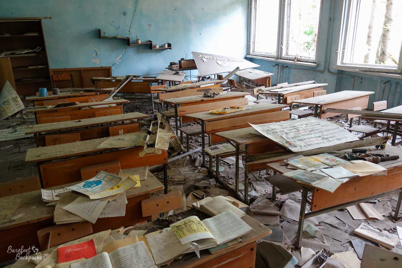 One of the classrooms in one of the schools in Pripyat. A series of wooden desks, wide enough for two people, are covered with fallen plaster, posters, and open books