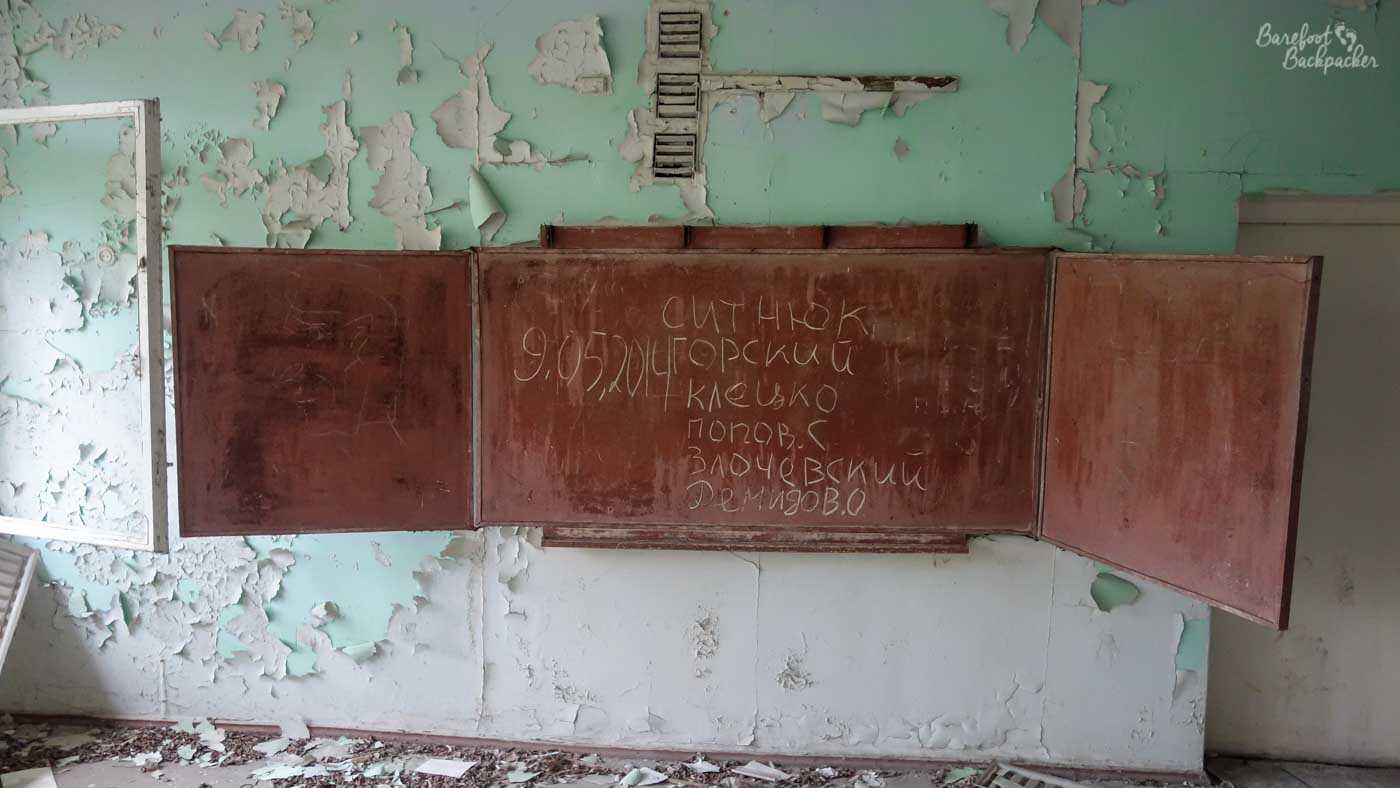 On a wall is a chalkboard or blackboard, one with doors that open out or close to cover it. They are open. On the board are written words in Cyrillic. The walls are peeling very badly.