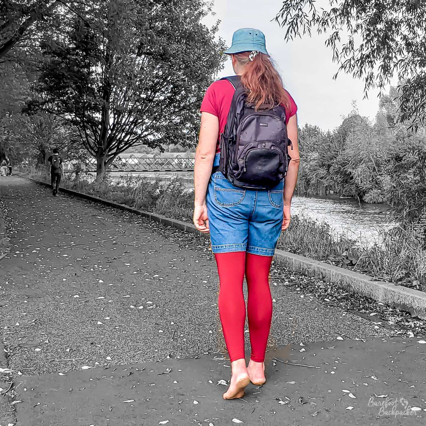 Someone, possibly a taller-than-average female, is walking away from the camera, on a solid path in a park. On the right, down a dip, is a river. In the background are trees. The person is wearing a hat, a backpack, a t-shirt, dungaree shorts, and leggings. She is barefoot, and has a ponytail. With regard to the image itself, they are in full colour (blue dungs and hat, red t-shirt and leggings), but the rest of the scenery - the trees, the path, the river, etc - has been rendered in black & white.