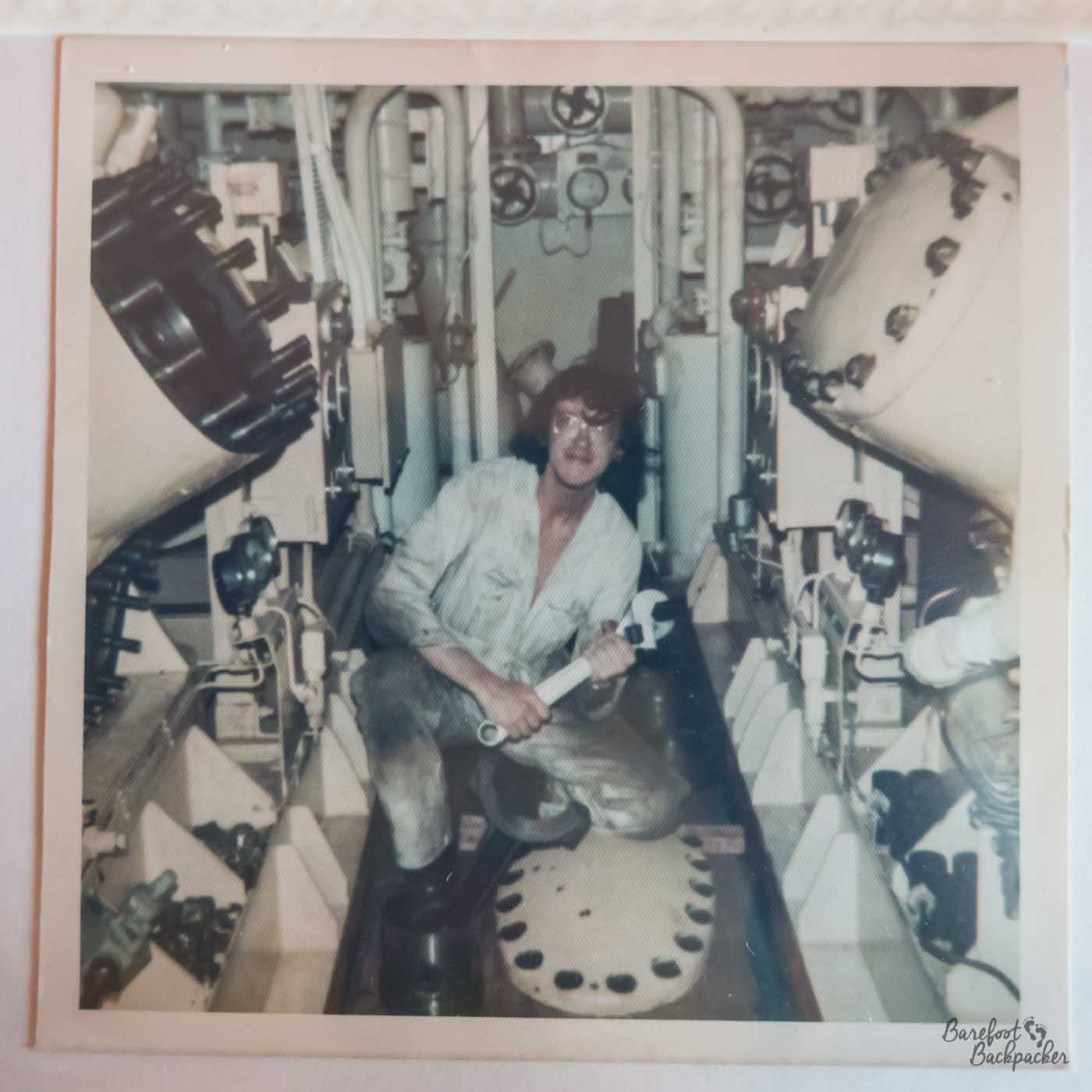 A photo taken of a previously-taken picture, possibly a polaroid, in a faded 1970s way. The older picture is of the inside of an engine room type environment. A man in a dirty boiler suit, boots, and glasses, is crouching down between some large pieces of engineering on either side of him. He is holding a large spanner or wrench.