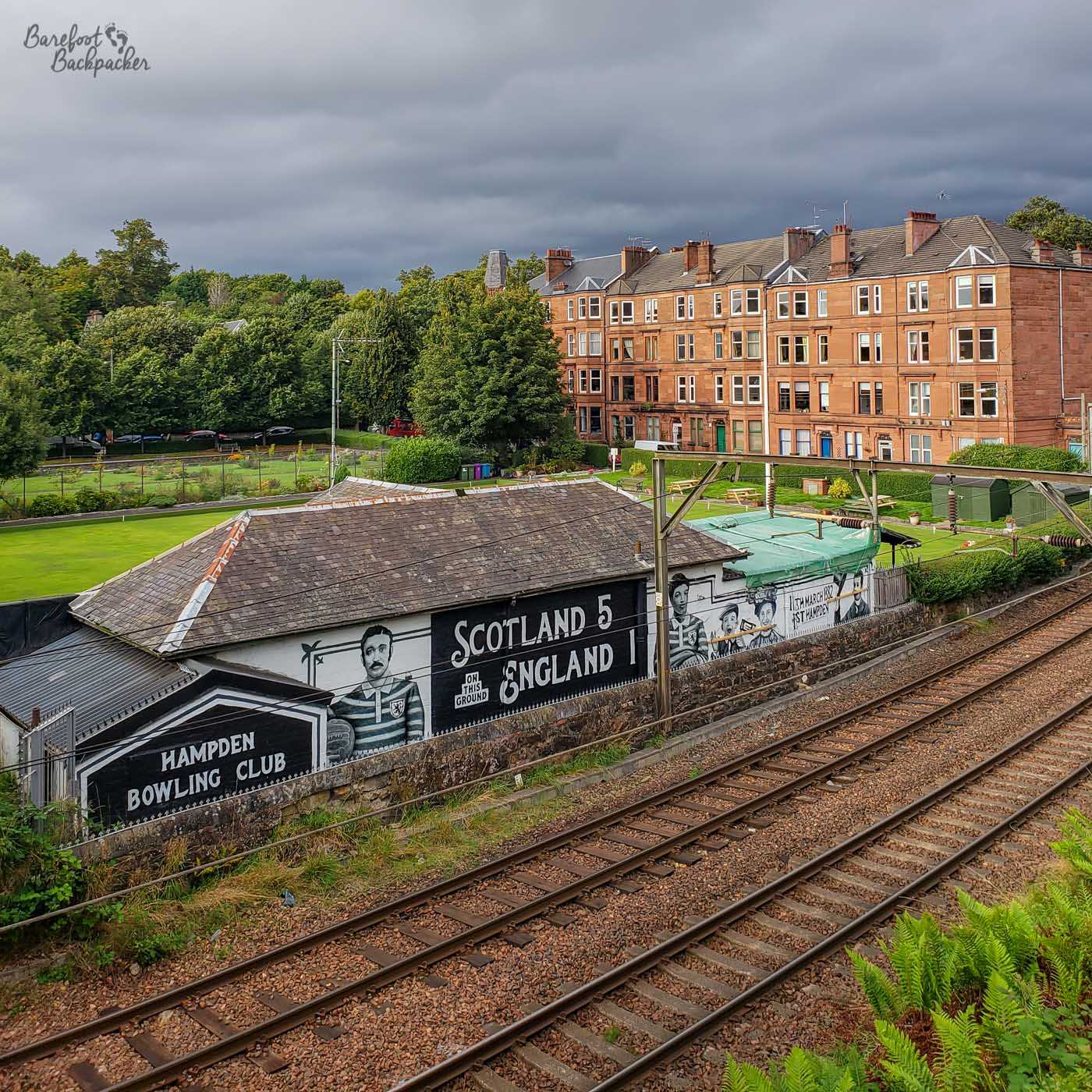 Shot from higher ground of a double-track railway line, behind which is a long, short building advertising itself on the roof as 'Hampden Bowling Club' but then several painted figures and the words 'Scotland 5 England 1'.