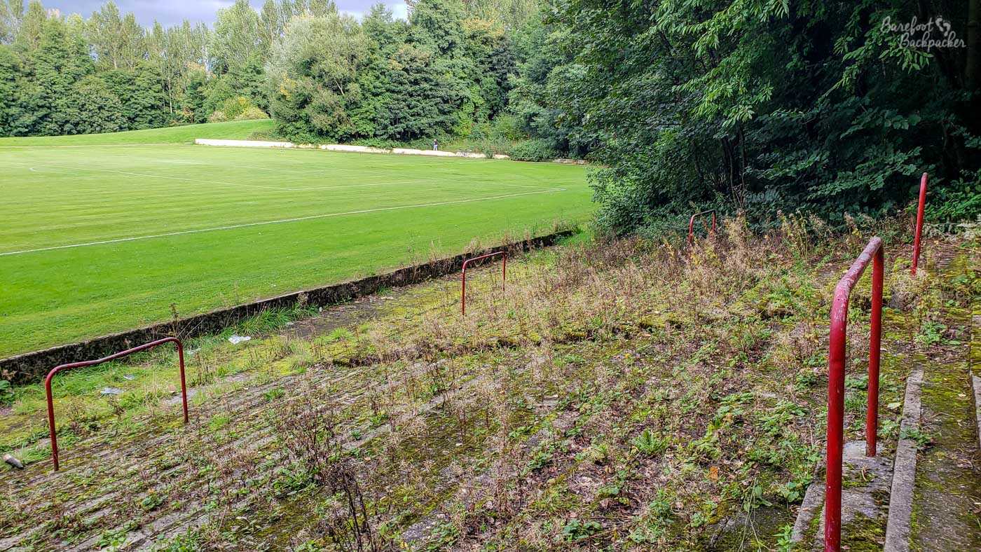 Terracing with metal barriers, the stone floor overgrown with weeds, The terracing slopes to a grassy pitch with a small wall in front. On the right, the run of the terracing is blocked by large bushy trees.