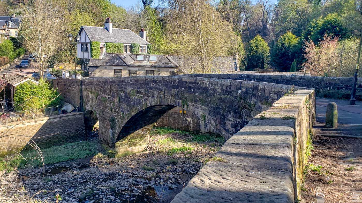 A single-arch stone bridge crosses a river. In the background are trees and old-looking houses.