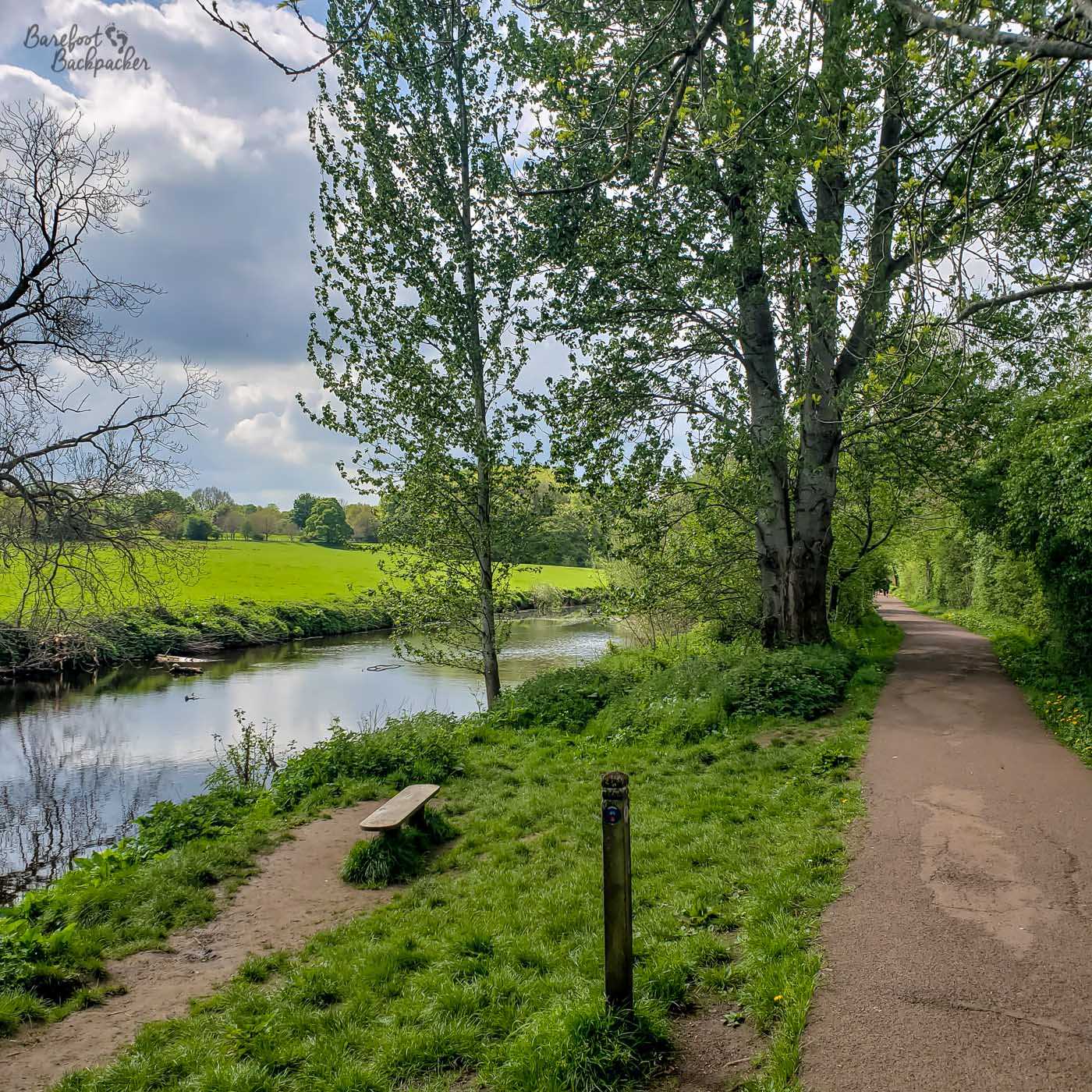 A path in the countryside, next to a river. There's trees between the path and the river, and the other side of the river is an empty field.