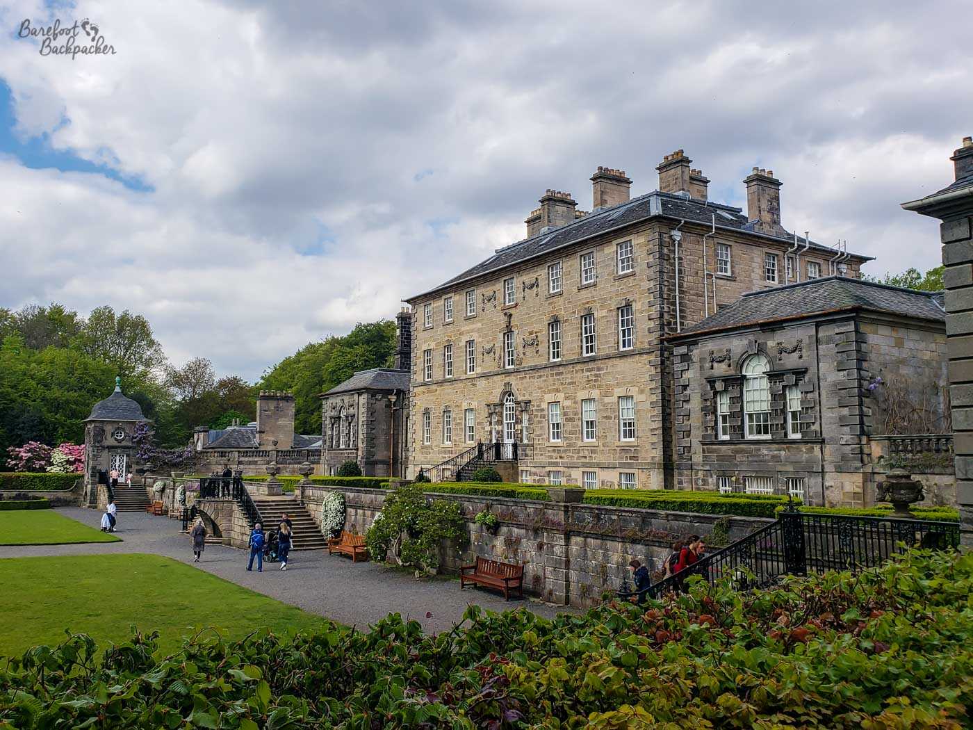 A large rectangular stone house with square windows, it stands on a raised embankment, and there's a wide stone staircase leading up to it. At the bottom and around the staircase are landscaped gardens.