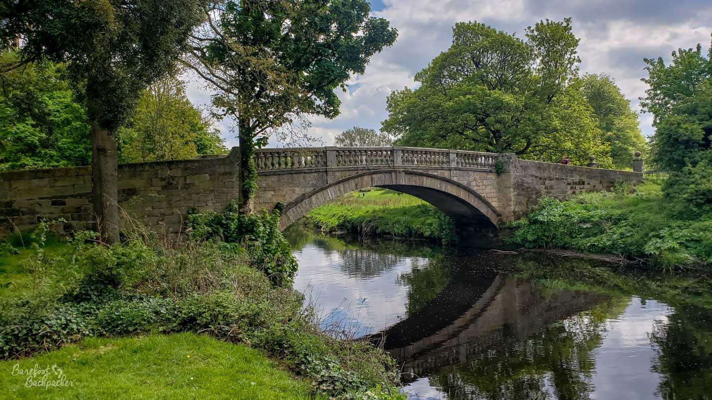 A stone single-arch bridge spans a river. Either side of the river is grass and trees.