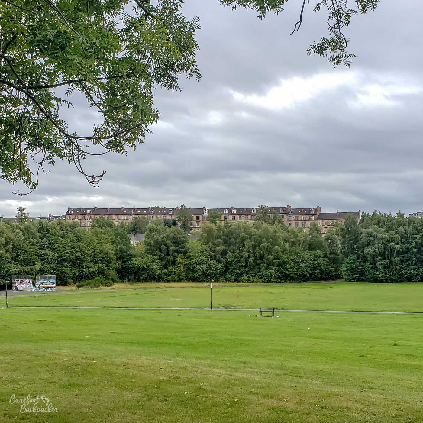 Shot overlooking a large plain grassy park, taken from up a small incline. There's a path just visible crossing from left to right, trees at the far end of the park, and tenements behind.