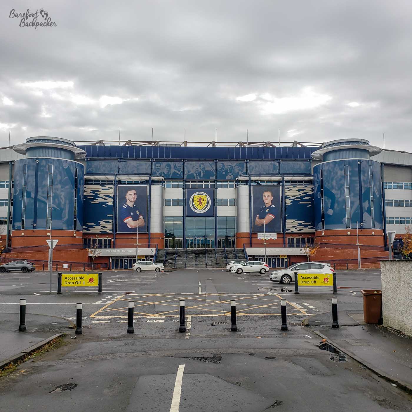 A car park, beyond which is a stone staircase leading up to a building. On either side of the portico are huge pictures of Scottish footballers and, to the sides of those, two large rounded columns/towers.
