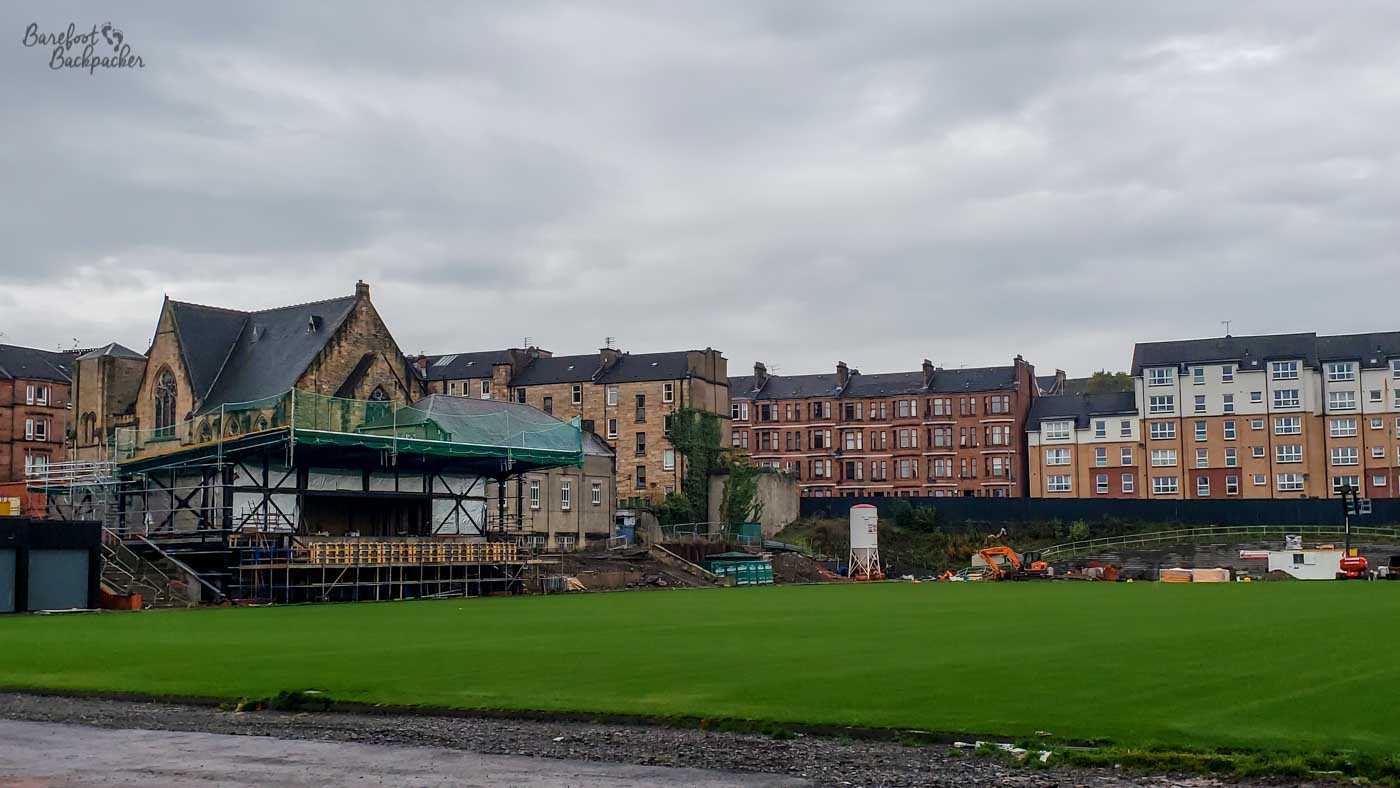 View inside a stadium under construction; the pitch is present but the stands are still being built. Aside from the framework of a small one to the left side, there is just banks of soil. There are tenements and a church behind.
