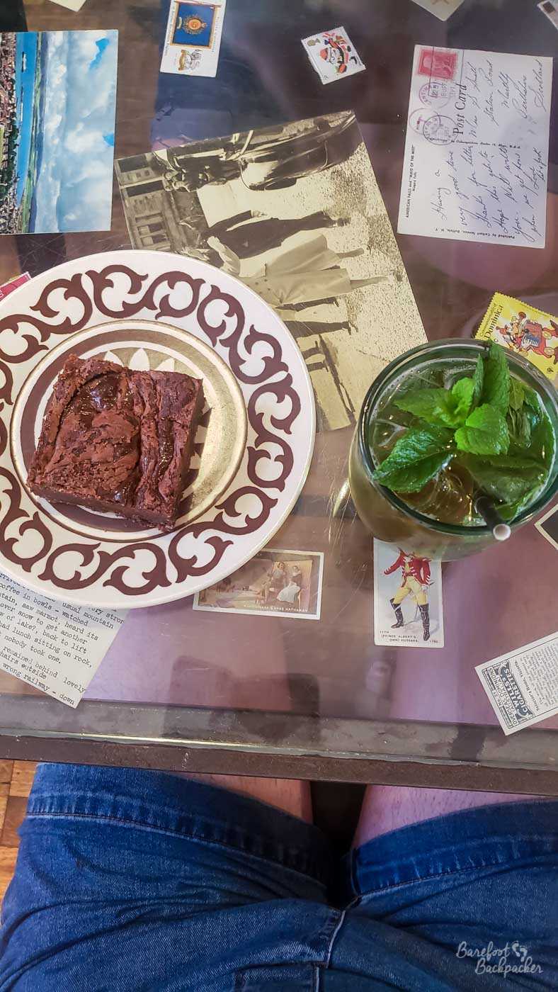 A shot taken looking down at a table in a cafe. The table is glass-topped and has stickers and cards between the frame and the glass. On the tabletop is a chocolate brownie and a glass filled with mint leaves.