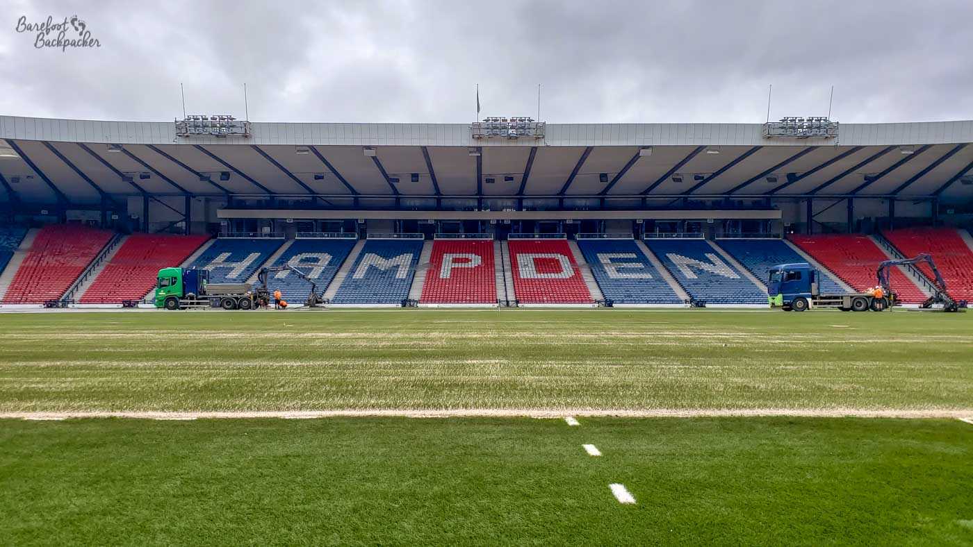 View inside an empty sports stadium, looking across the pitch to a stand where the seating spells out the word 'Hampden'.
