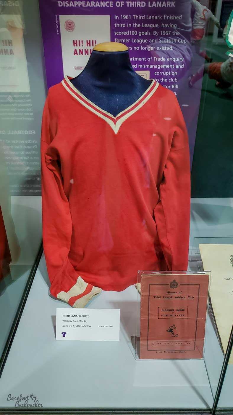 A red long-sleeved football jersey with white trim is on display inside a perspex box.