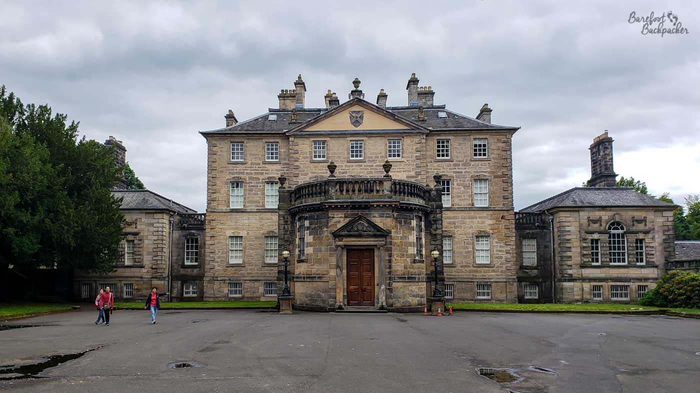 A large rectangular stone house with square windows and a semi-circular entrance portico stands behind what appears to be an empty car park.