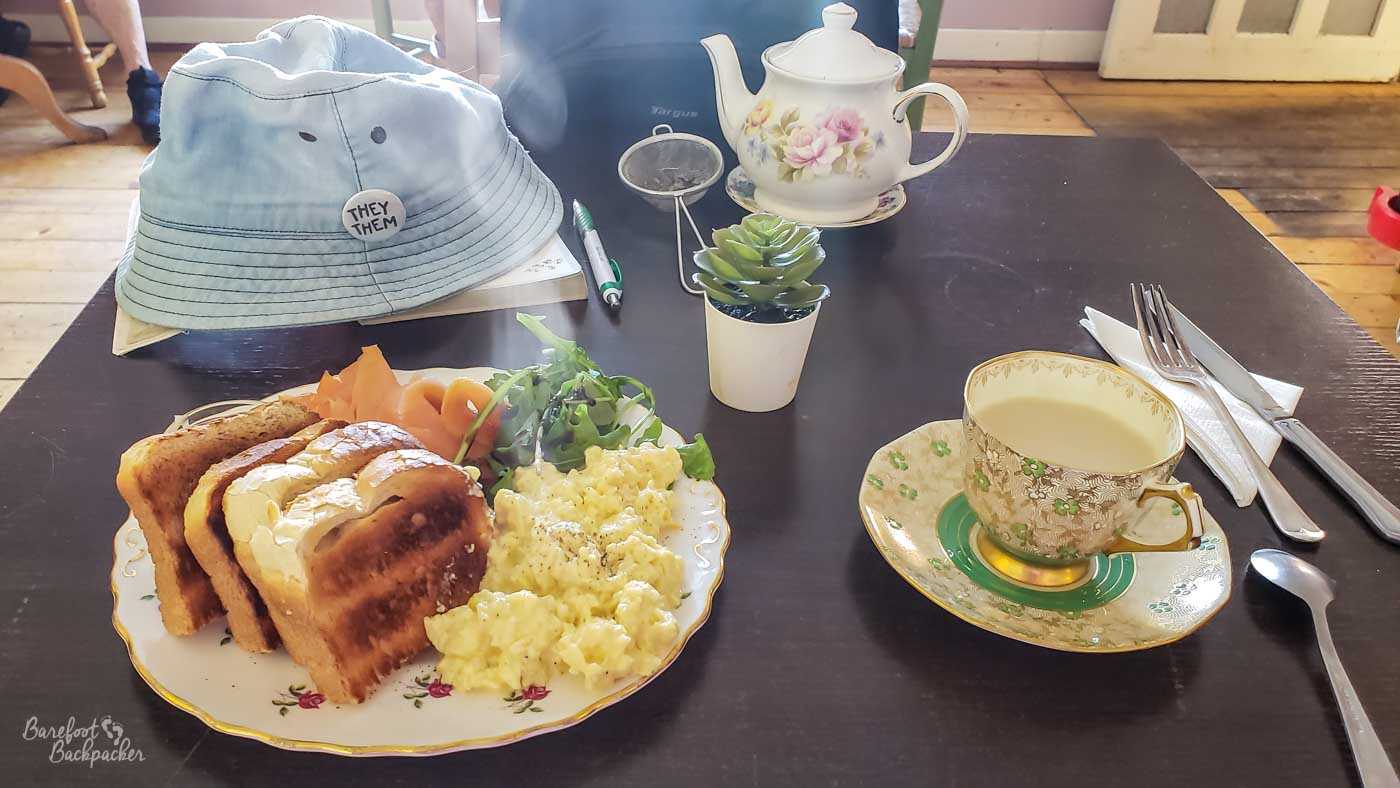 Floral-patterned teacup, saucers, and teapot, on a table along with a teastrainer and a small potted plant, and a plate with toast, egg, salmon, and green leaves on it.
