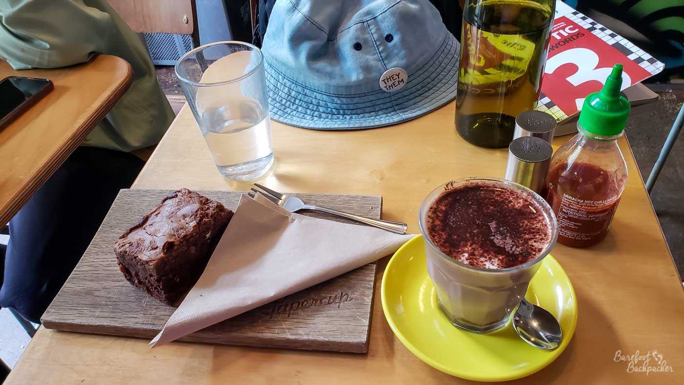 Chocolate Brownie, Hot Chocolate, salt, pepper, chilli sauce, a glass and bottle of water, a blue hat, and a book of crosswords are on a flat single piece of wood topping a table.