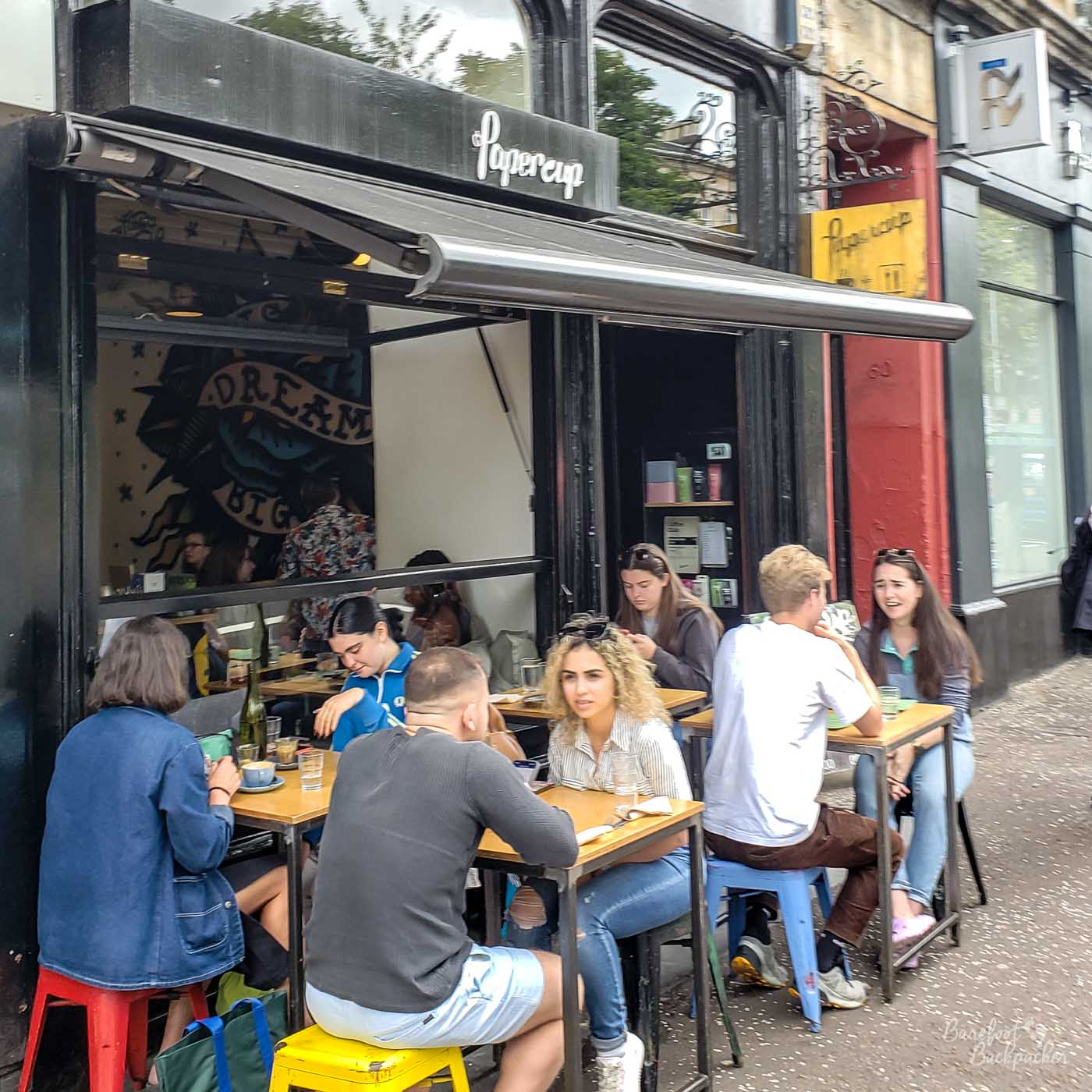 Four tables, each with two people on, on a street outside the cafe's front. The tables are underneath a canopy.