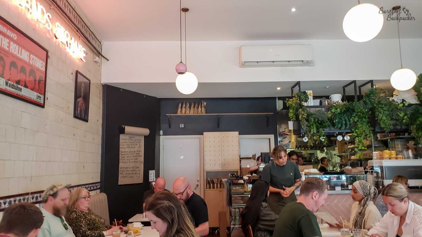 Wideshot of the inside of a cafe, with lots of people sat at tables. On the left wall, tiled, are a couple of 1960s-era posters. On the right, by the serving counter, is foliage.