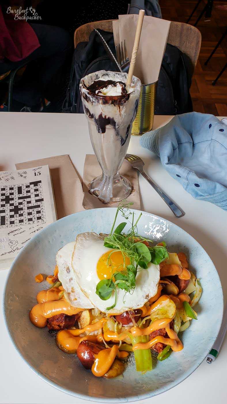 Sausage and egg salad, with greenery, a chocolate milkshake in a large glass, and a book of crosswords, are on a table in a cafe.