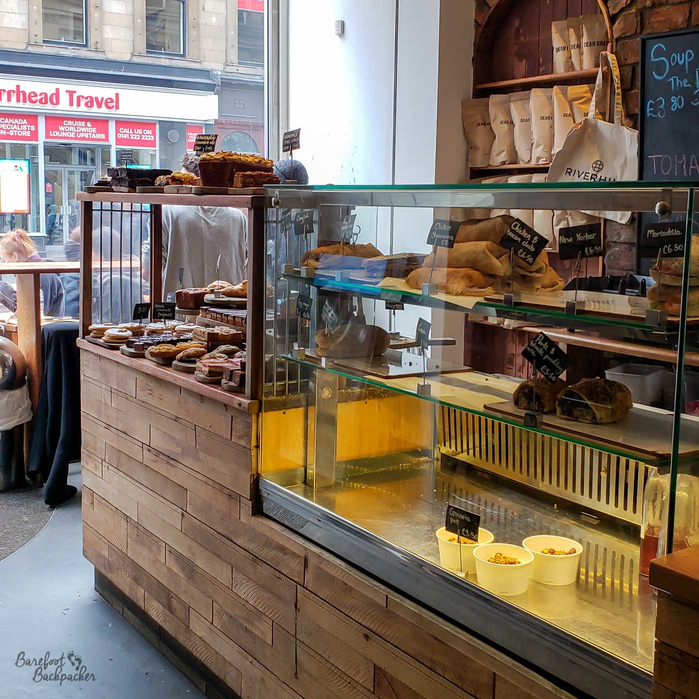 A counter with display sections in a cafe. It's wooden-panel-fronted, with cakes and bread on the left, and sandwiches and pies on the right, behind a perspex screen.