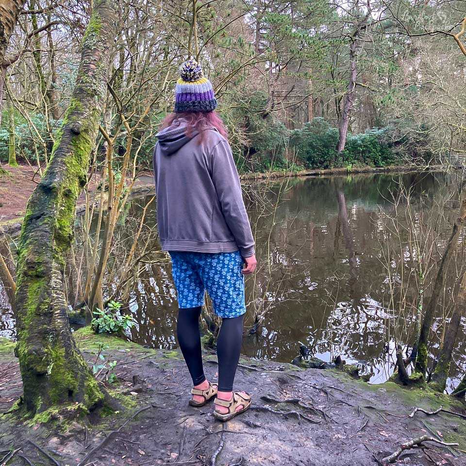 An enby stands on a batch of muddy earth, looking out over a small lake surrounded by thick trees.