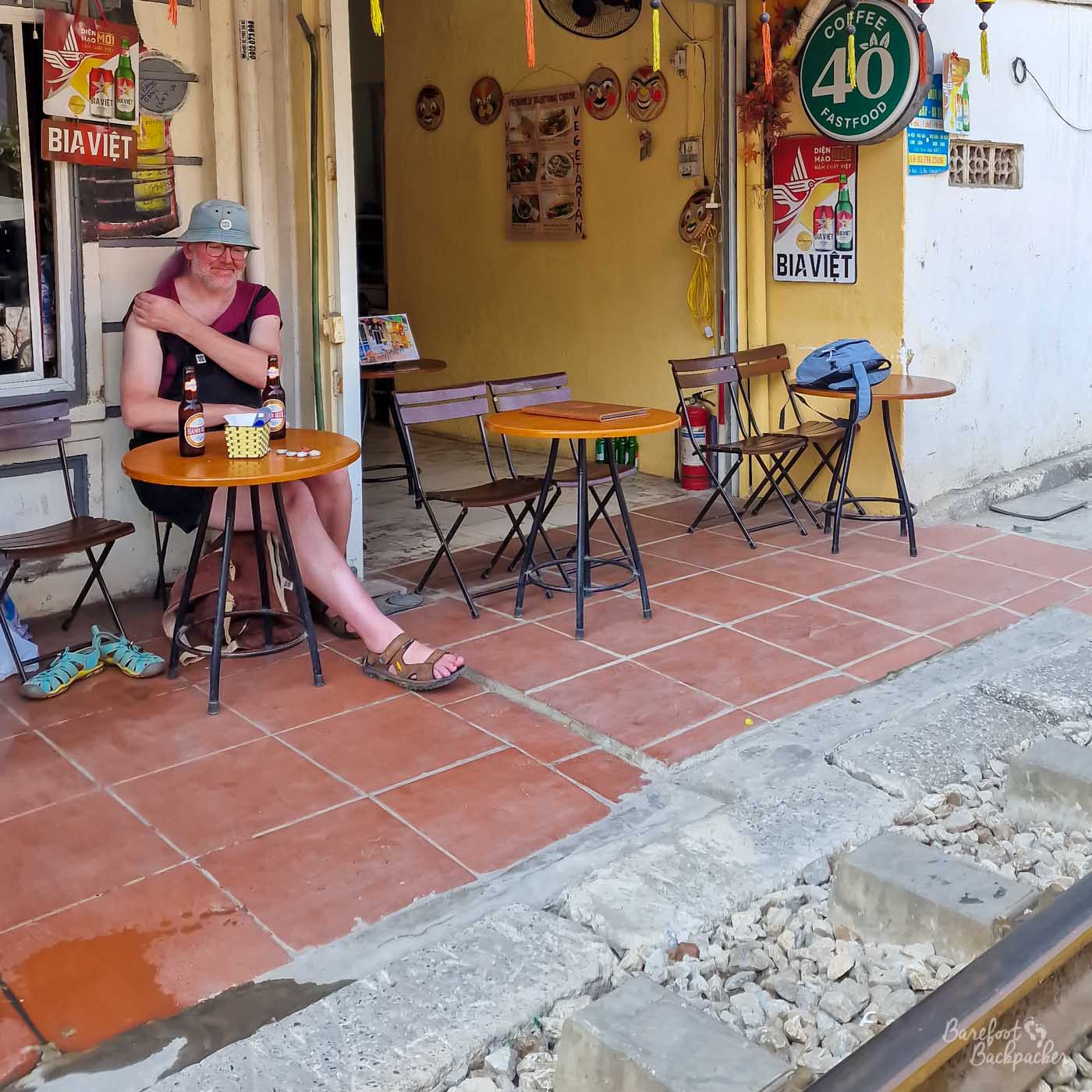 Someone is sat on a stool by a small table in front of a cafe. The walls of the cafe are plain and covered with posters for Vietnamese beer.