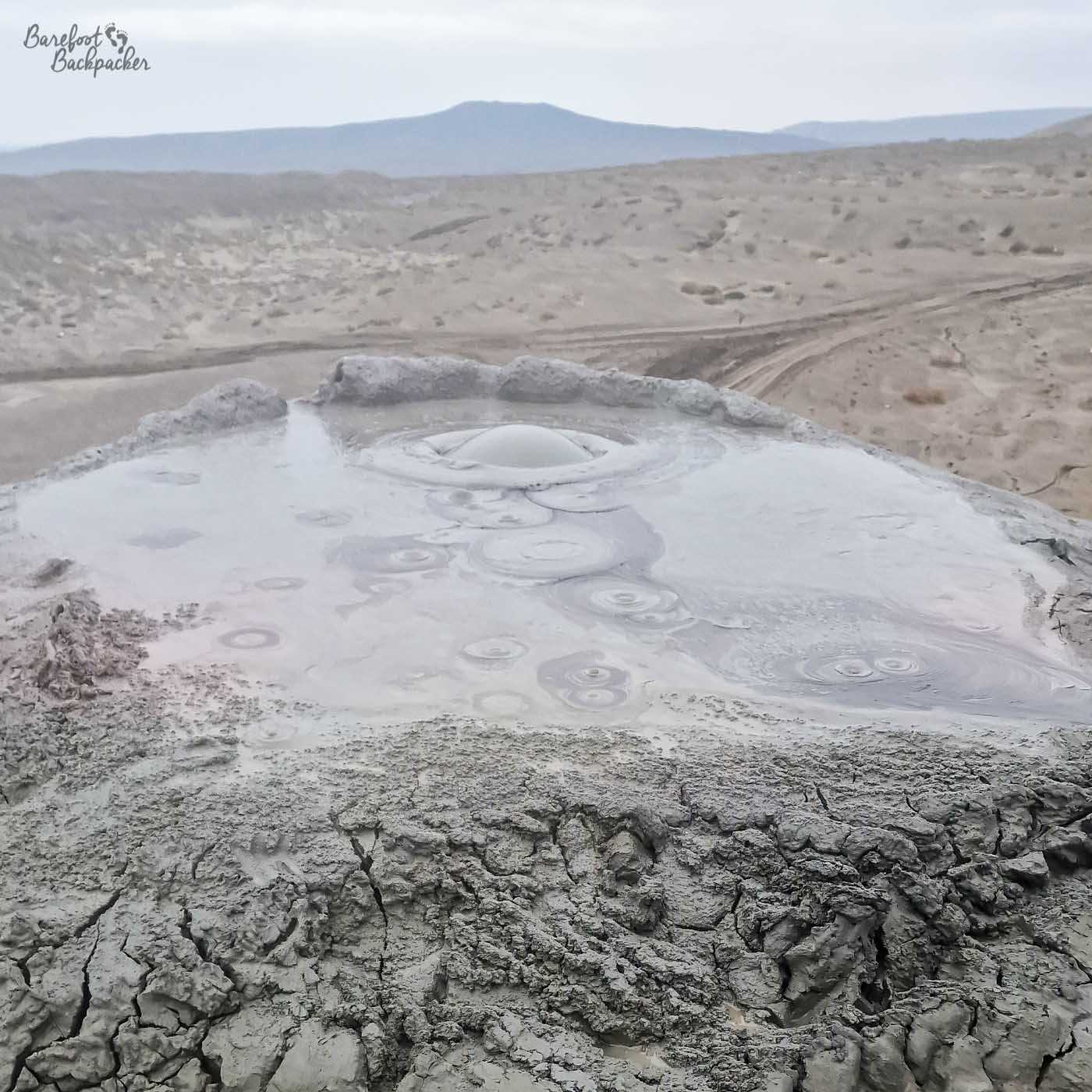The rounded mound on top of a small hill. On it is a pool of grey, bubbling. mud. The landscape below in the background is quite flat and desolate.