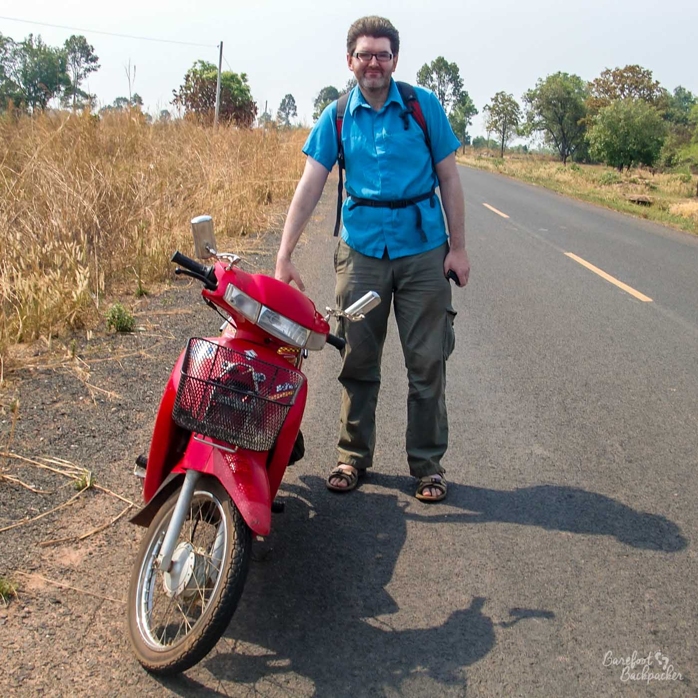 A male backpacker stands next to a motorbike, on an otherwise empty road going through shrubland.