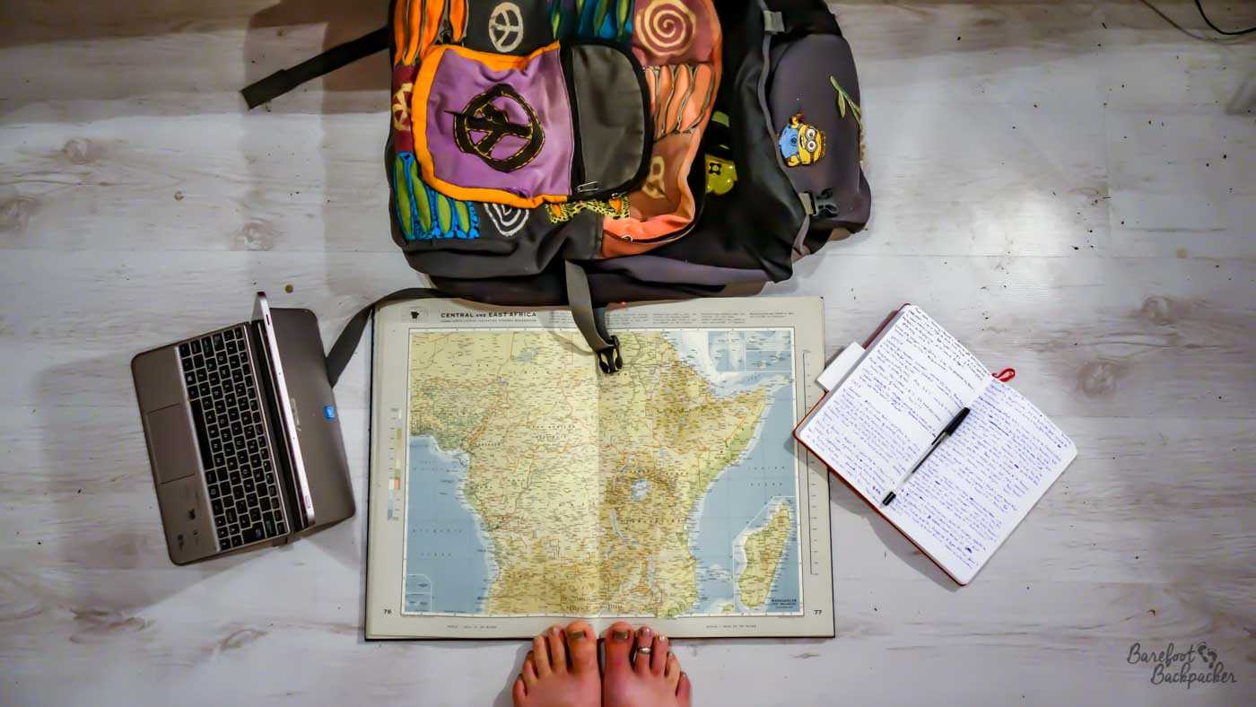 A top-down shot reveals a travel-themed scene on a light, weathered wooden floor. In the upper part of the frame, a multicoloured backpack with various patch designs leans slightly to the right. Beneath it, a map of Central and East Africa is spread open, its detailed topography and borders visible. To the left of the map is a silver laptop, partially open, displaying its keyboard. A red-covered notebook lies to the right of the map, filled with handwritten notes in blue ink and a pen resting on its pages. Near the bottom of the image, a pair of bare feet with painted toenails are visible, adding a personal touch to the scene.