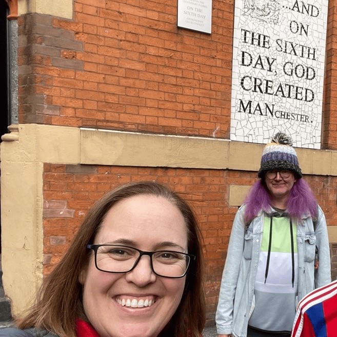 Two people in front of a brick wall with a sign on that says 'on the sixth day God created MANchester.