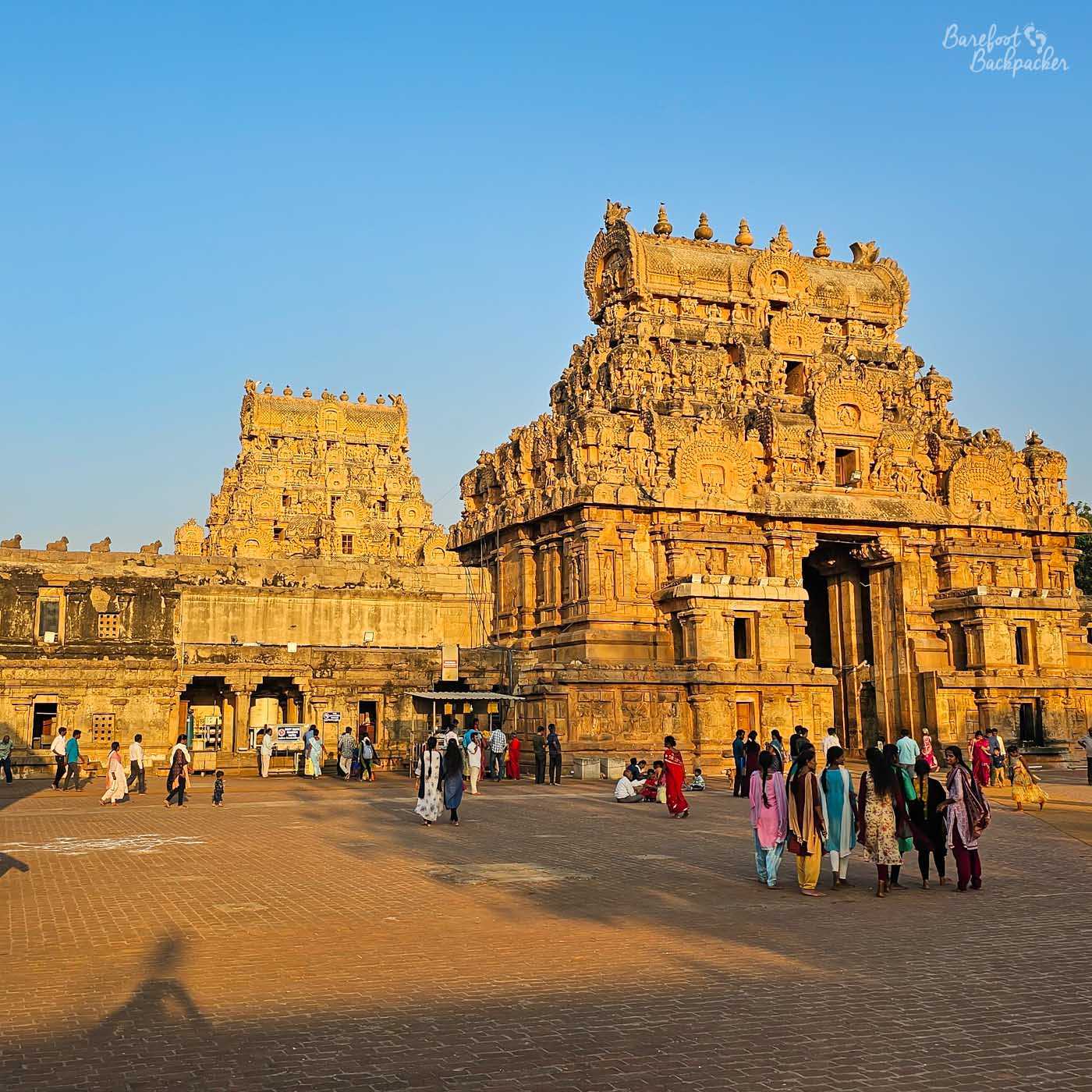 An exterior shot of the Brihadeeswarar Temple in Thanjavur, India. The towering temple, constructed of sandstone, is bathed in sunlight. The temple complex features two prominent gopurams (towers) adorned with intricate carvings. The foreground is a large paved courtyard filled with people.